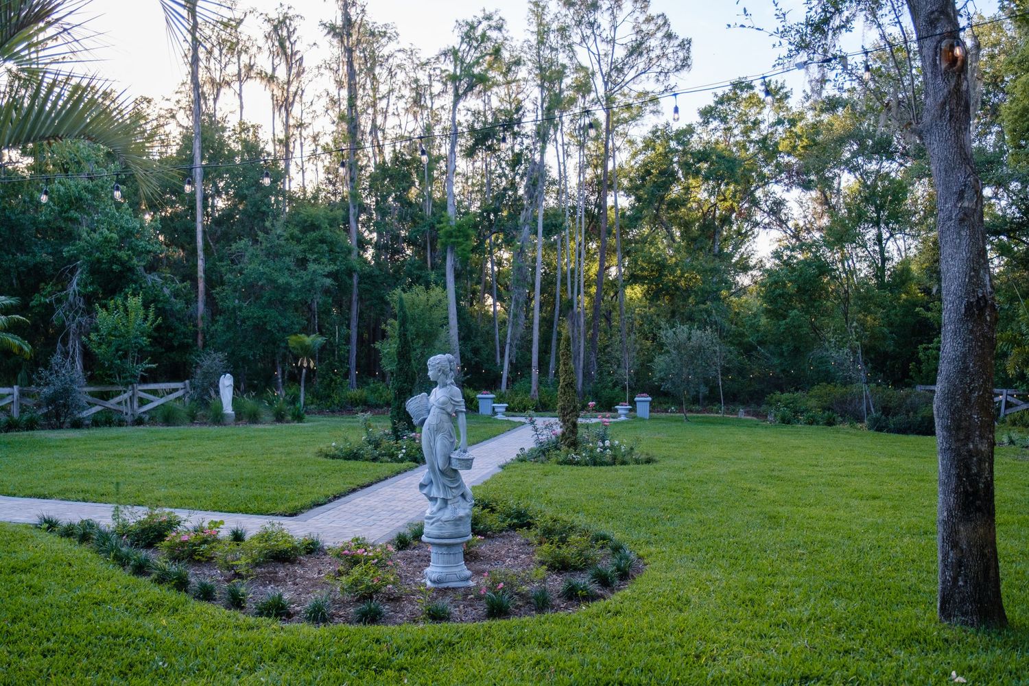 A classic garden statue in a landscaped circular bed surrounded by tall trees and manicured lawn at dusk.
