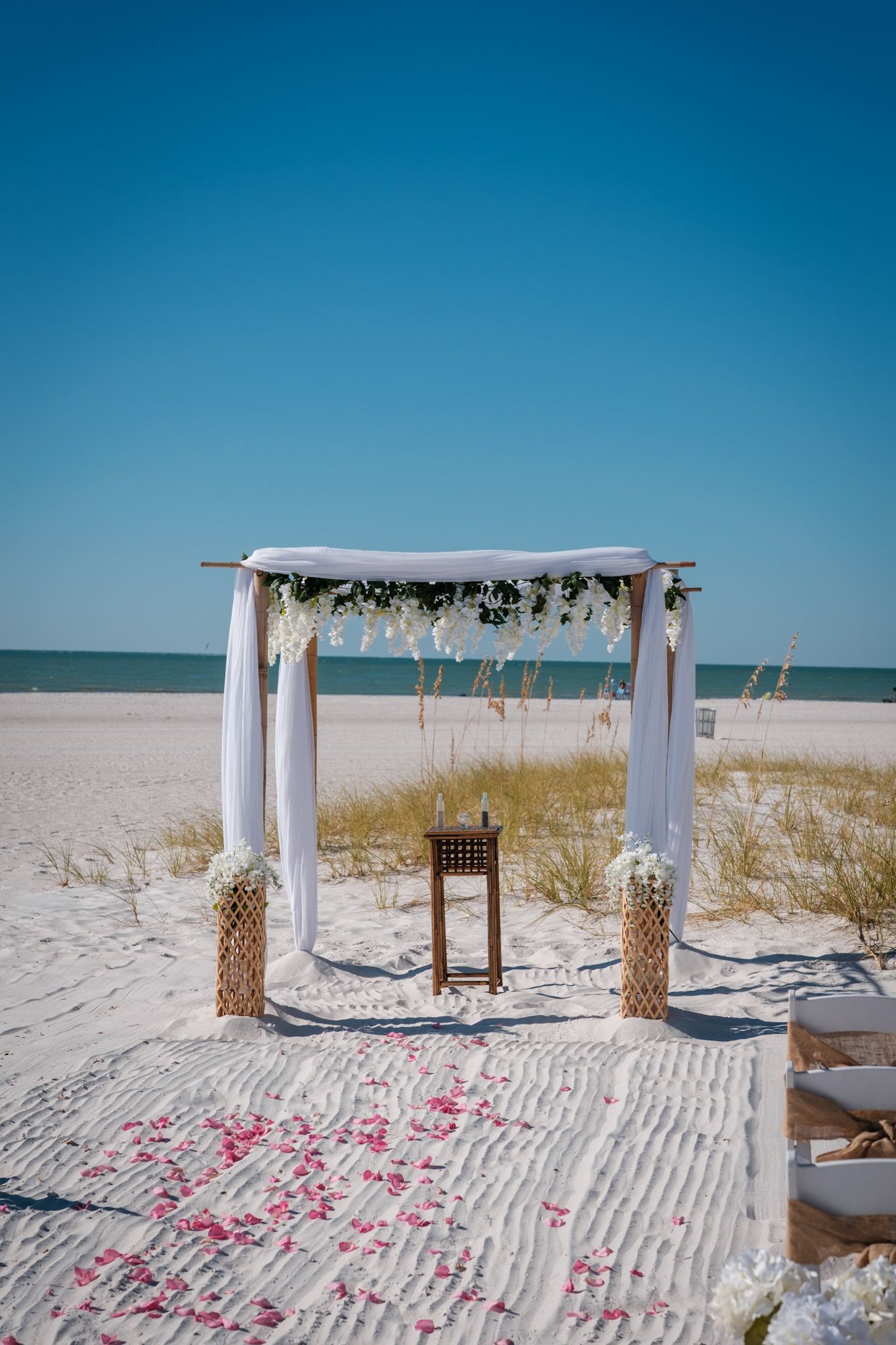 Beach wedding ceremony setup with white canopy arch decorated with flowers against bright blue sky.