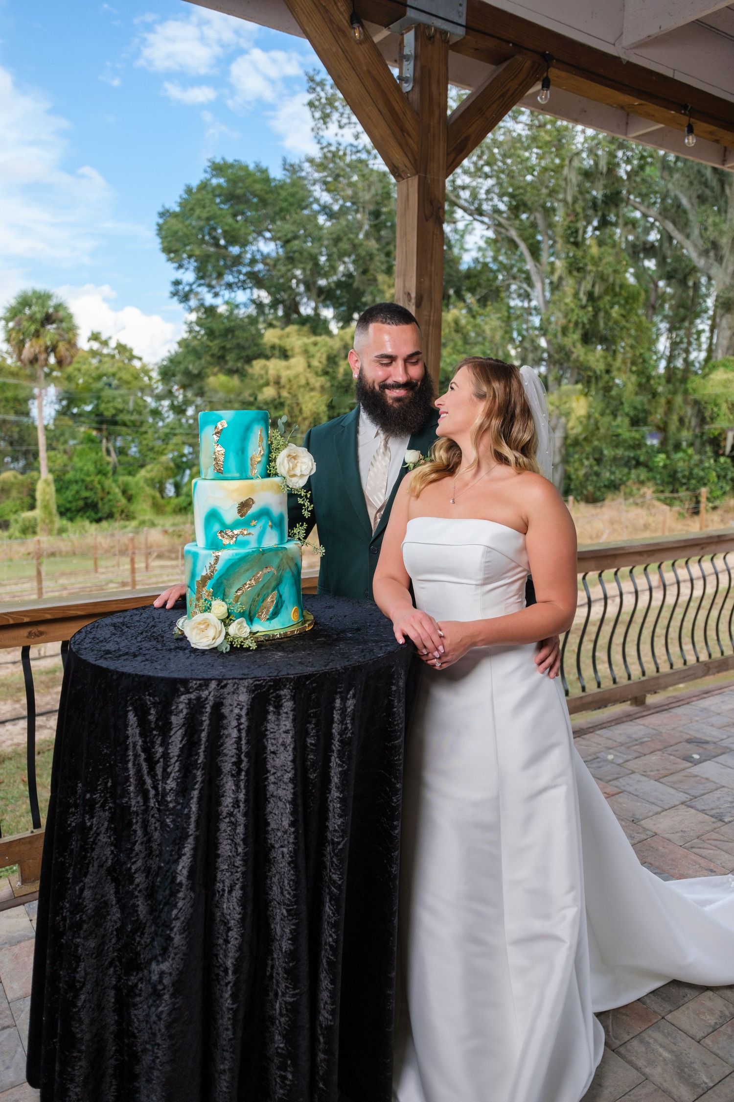 A couple stands near a turquoise and black wedding cake on a covered patio during their celebration.