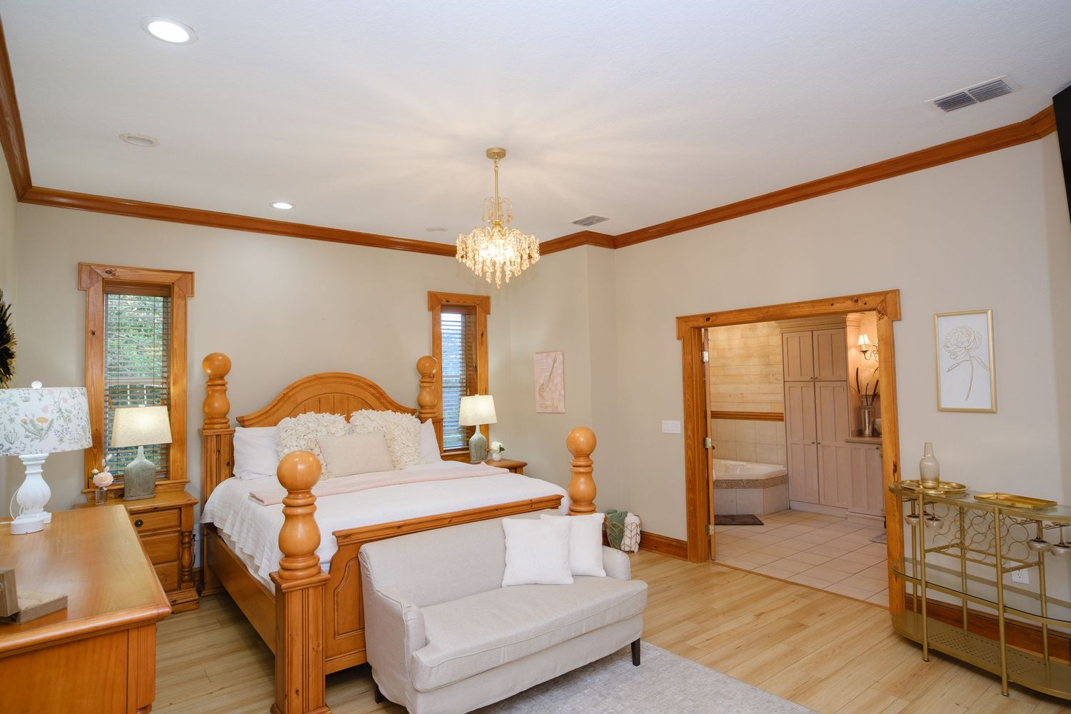 Spacious master bedroom with pine furniture, crystal chandelier, and natural hardwood floors bathed in sunlight.