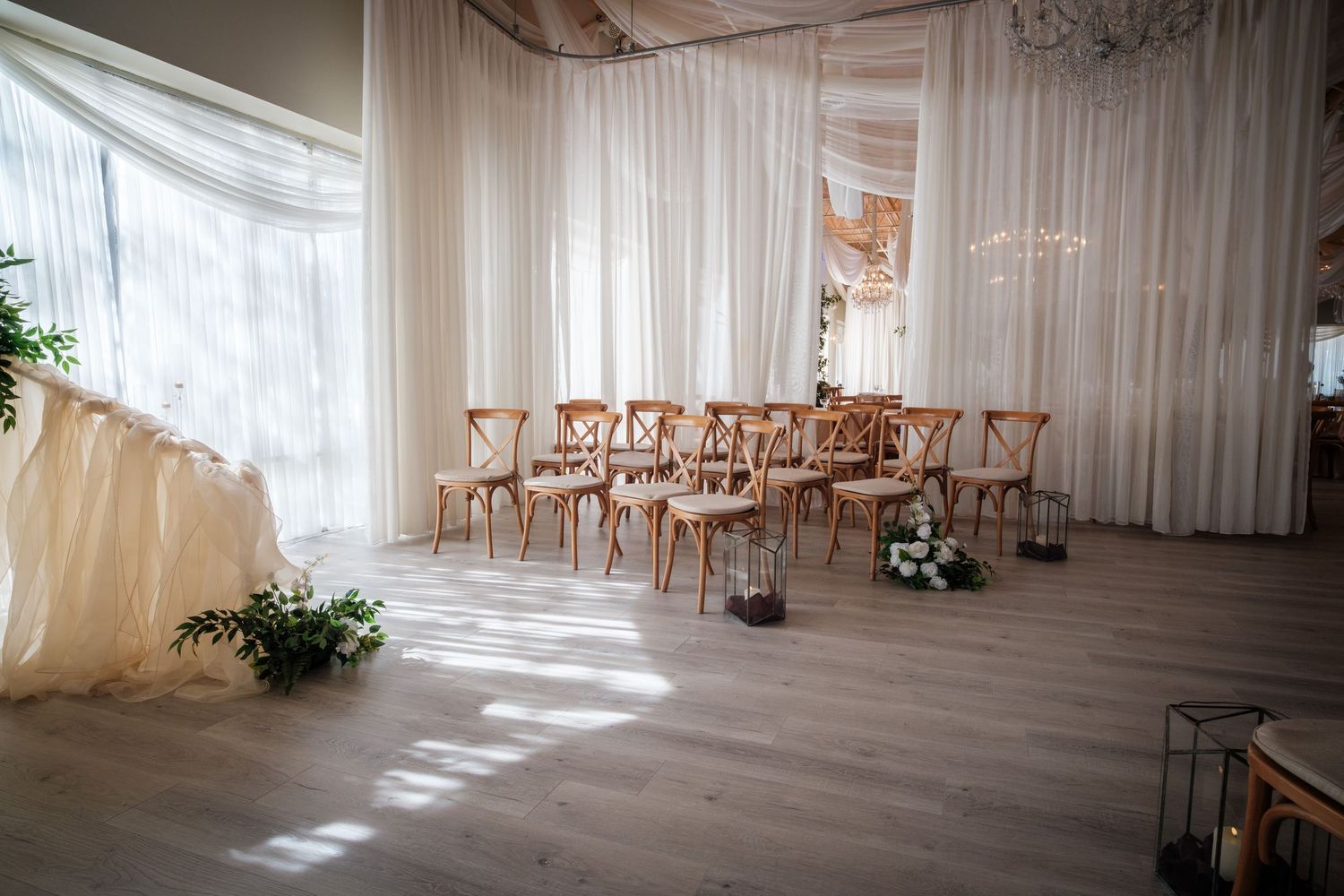Elegant wedding ceremony setup with wooden cross-back chairs, white draping, and floral arrangements in a bright venue.