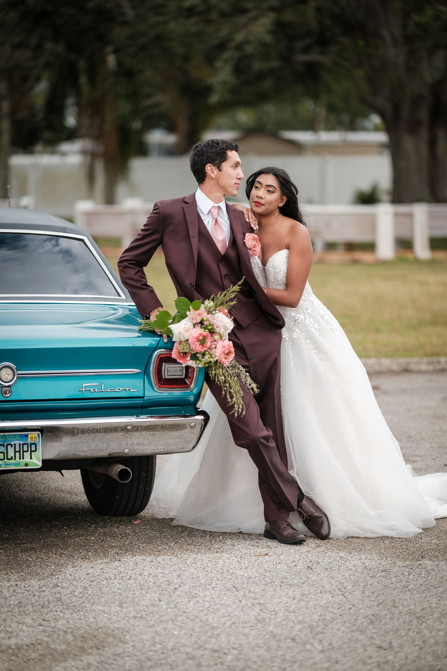 Newlywed couple poses with vintage turquoise car while holding pink floral bouquet at outdoor wedding venue.