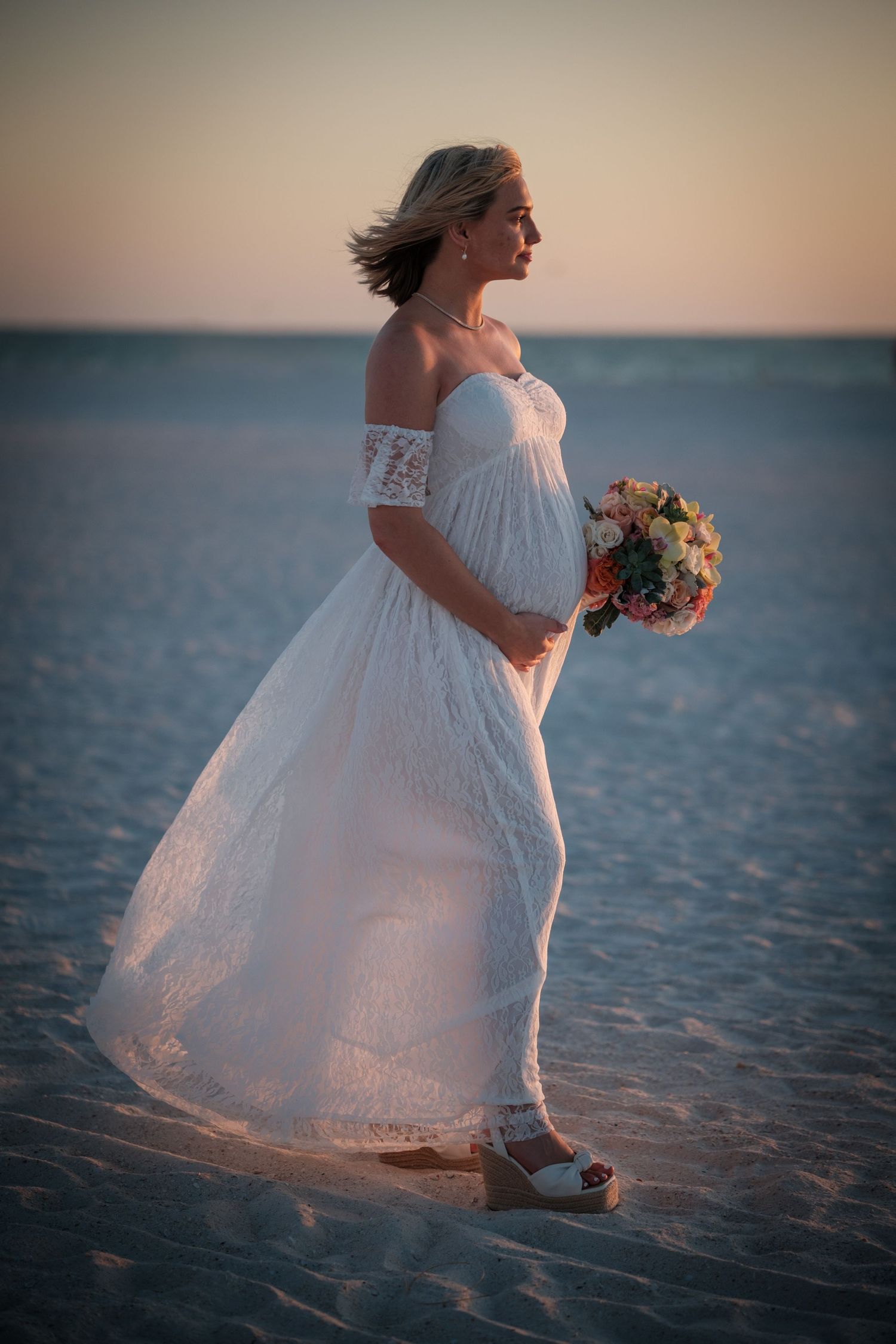 Pregnant bride in white off-shoulder lace gown holds bouquet on sandy beach at sunset.