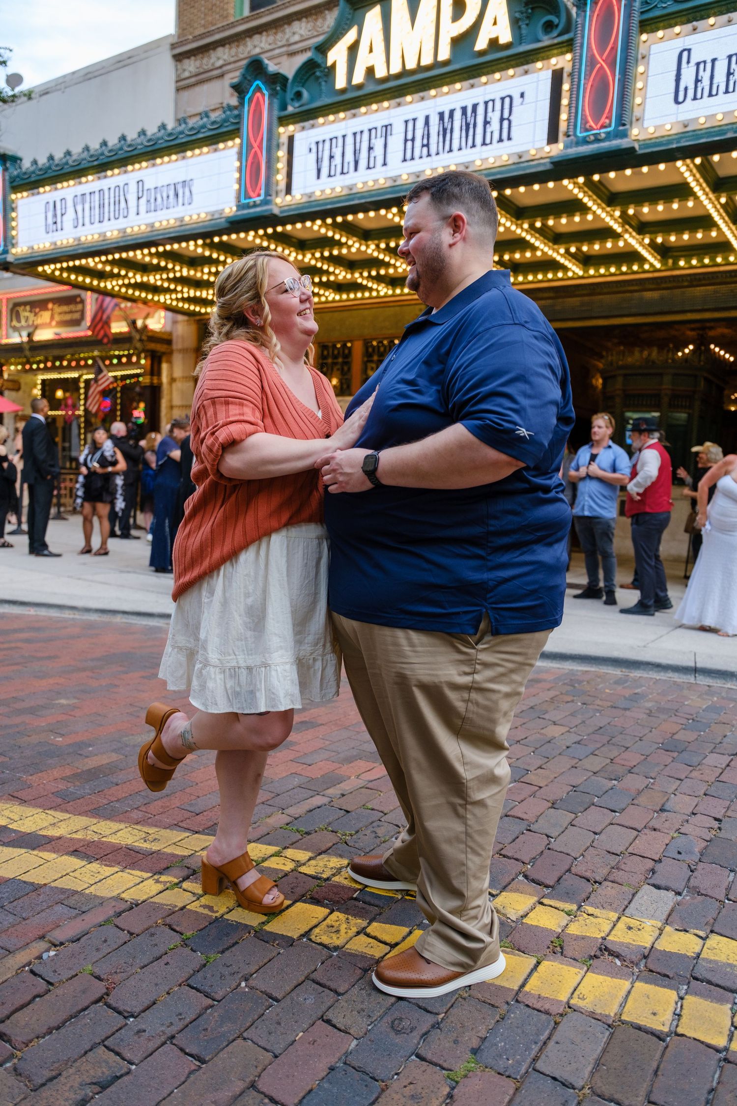 A couple dances together on a brick sidewalk in front of illuminated theater marquees in Tampa's entertainment district.