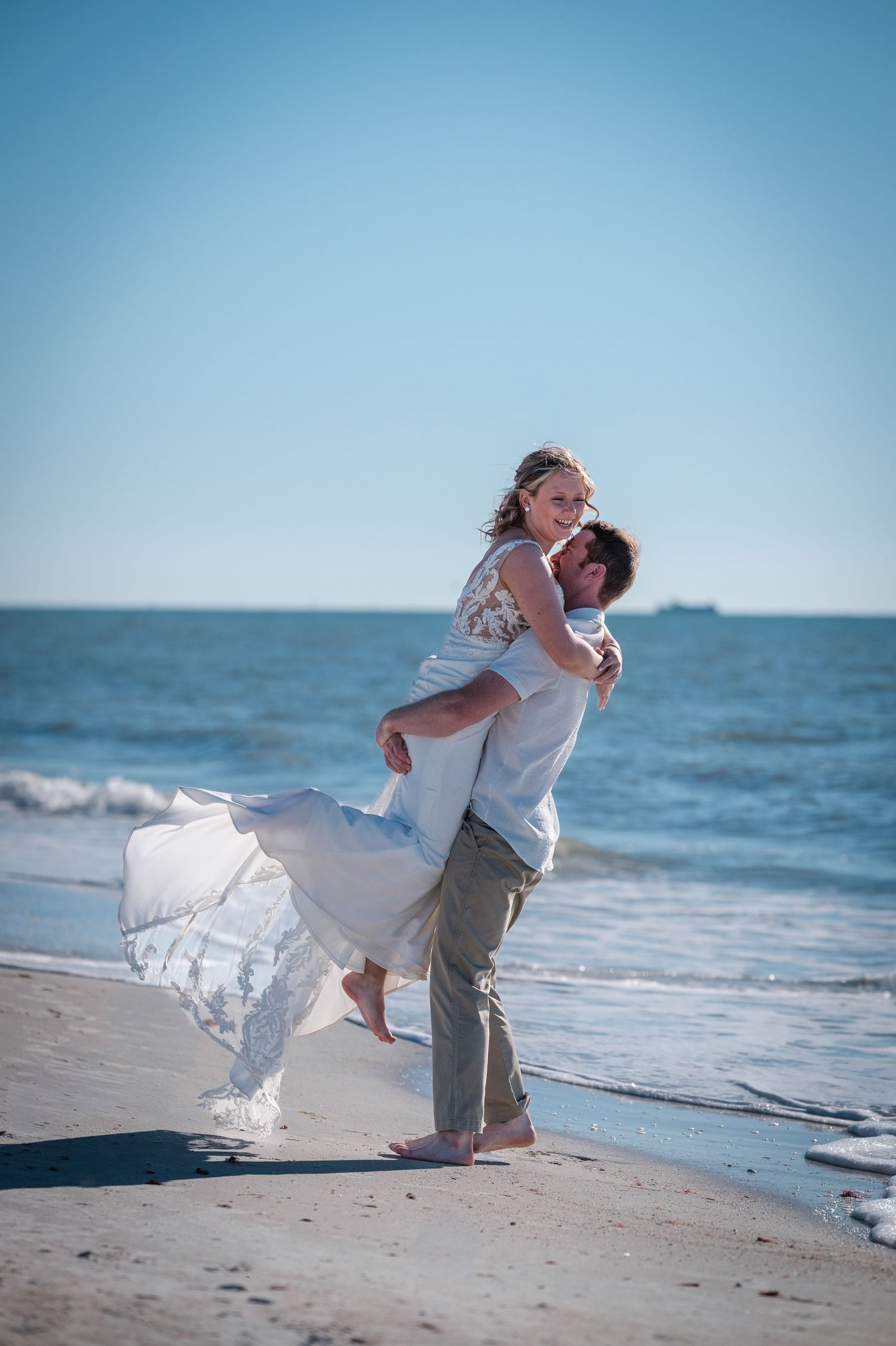 A romantic couple in wedding attire share an intimate dance pose on a sunny beach by the ocean.