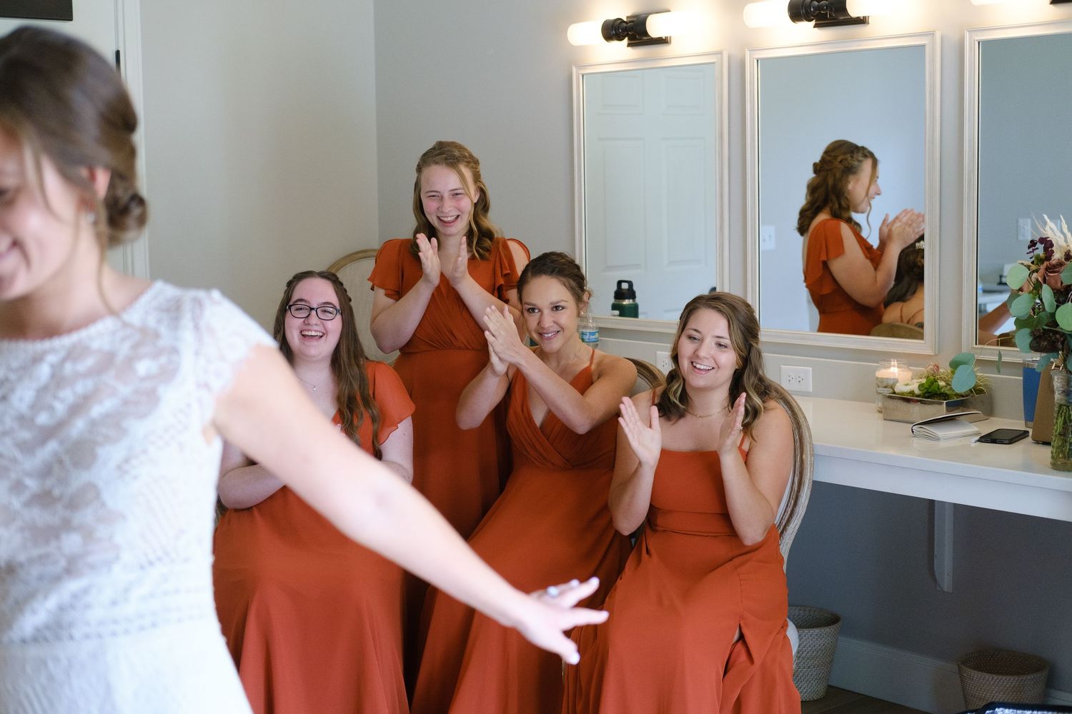 Bridesmaids in burnt orange dresses react with joy in a dressing room with illuminated mirrors.