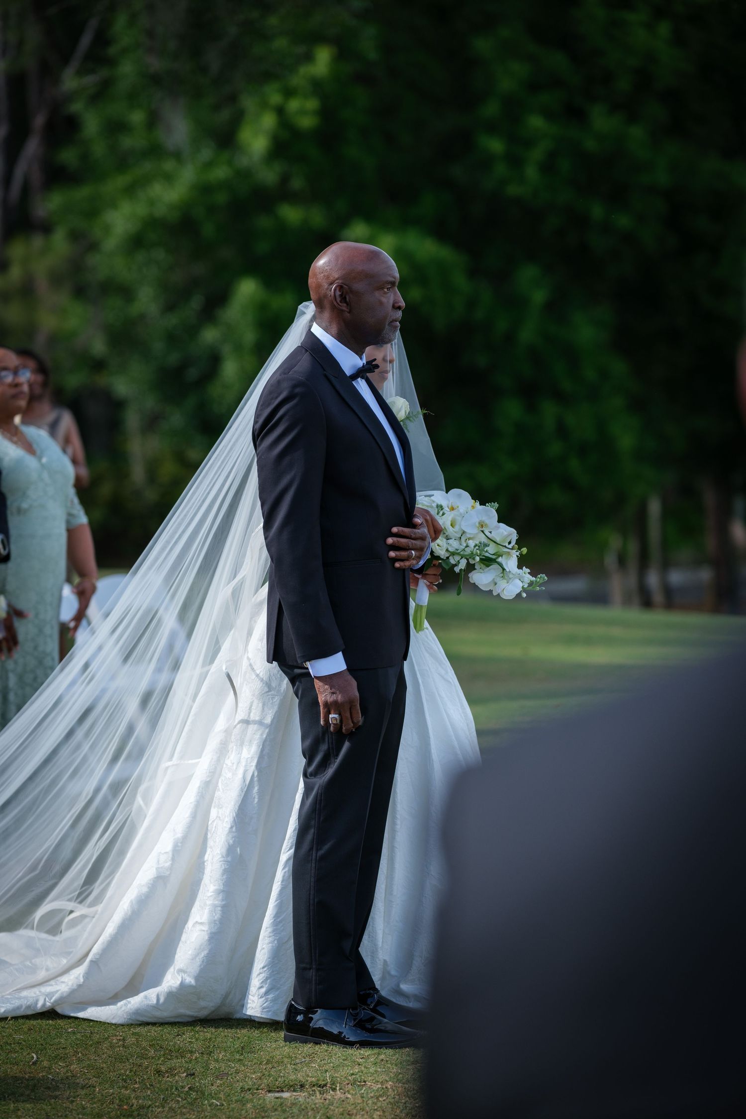 Groom in black tuxedo walks bride in white gown and veil down outdoor aisle holding bouquet.