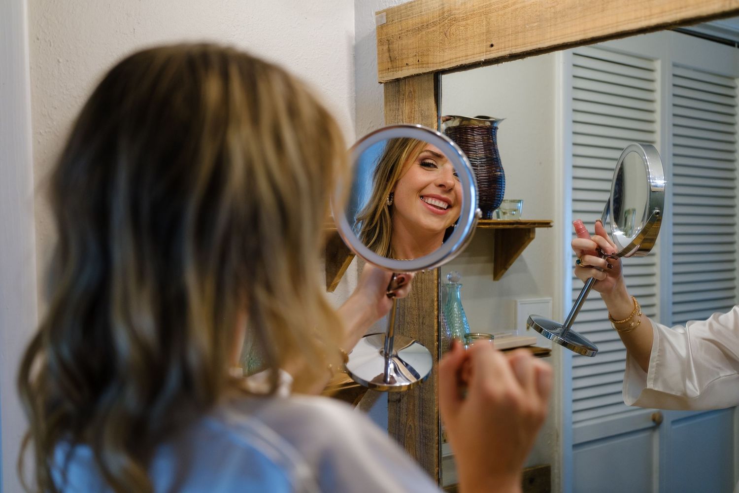 A woman with long blonde hair applies makeup while looking in a bathroom mirror.