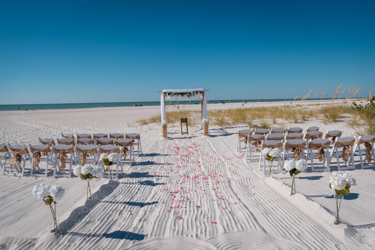 Aerial view of beach wedding setup with white chairs and rose petals scattered on wooden boardwalk by the ocean.