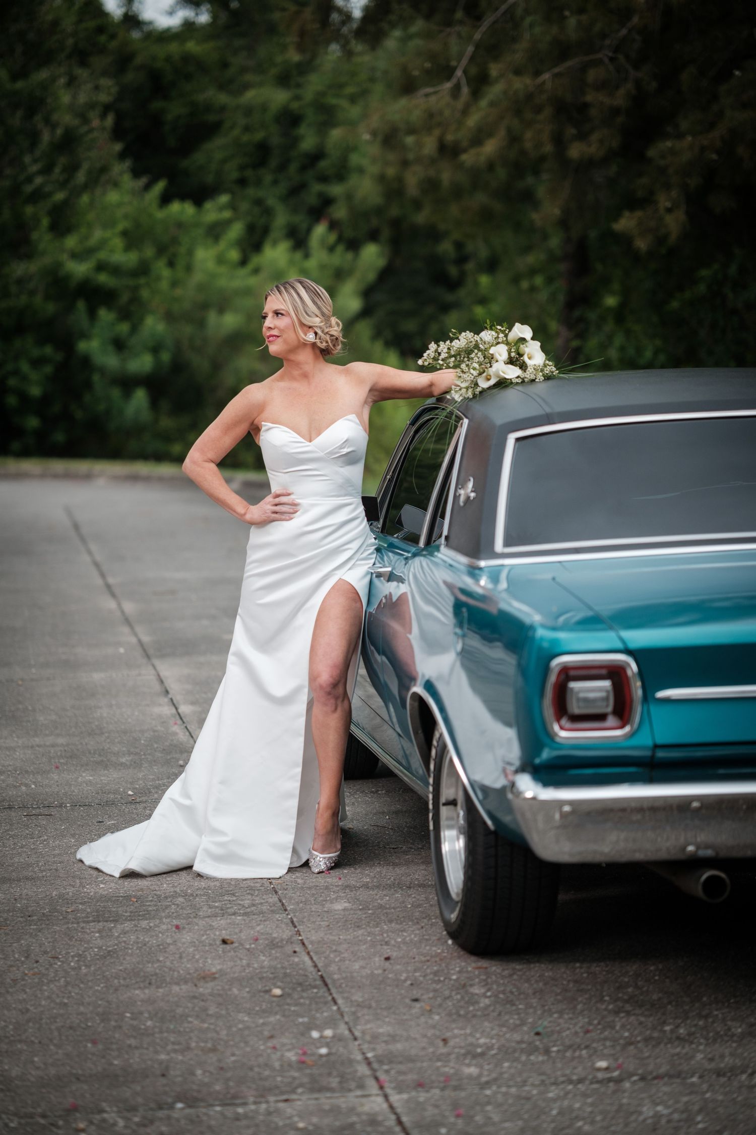 Bride in white satin gown with slit poses beside vintage teal car holding bouquet outdoors.