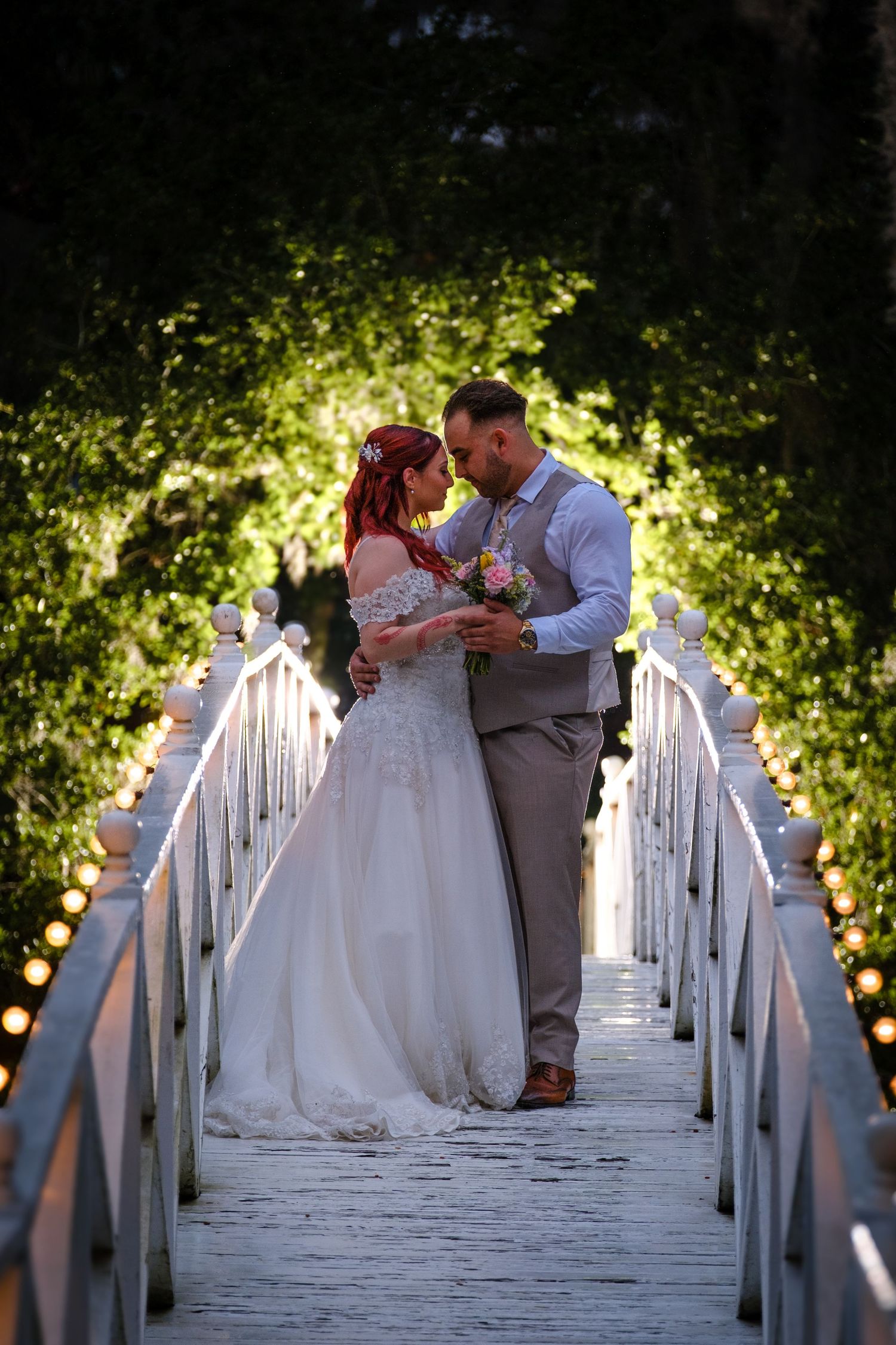 A couple shares an intimate moment on a white wooden bridge surrounded by glowing greenery and fairy lights at dusk.