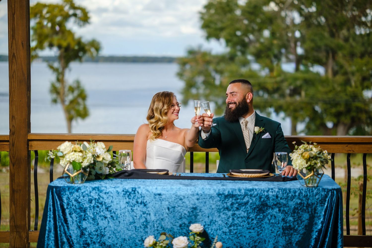 Wedding reception table with blue velvet tablecloth and glassware set against waterfront view on outdoor deck.