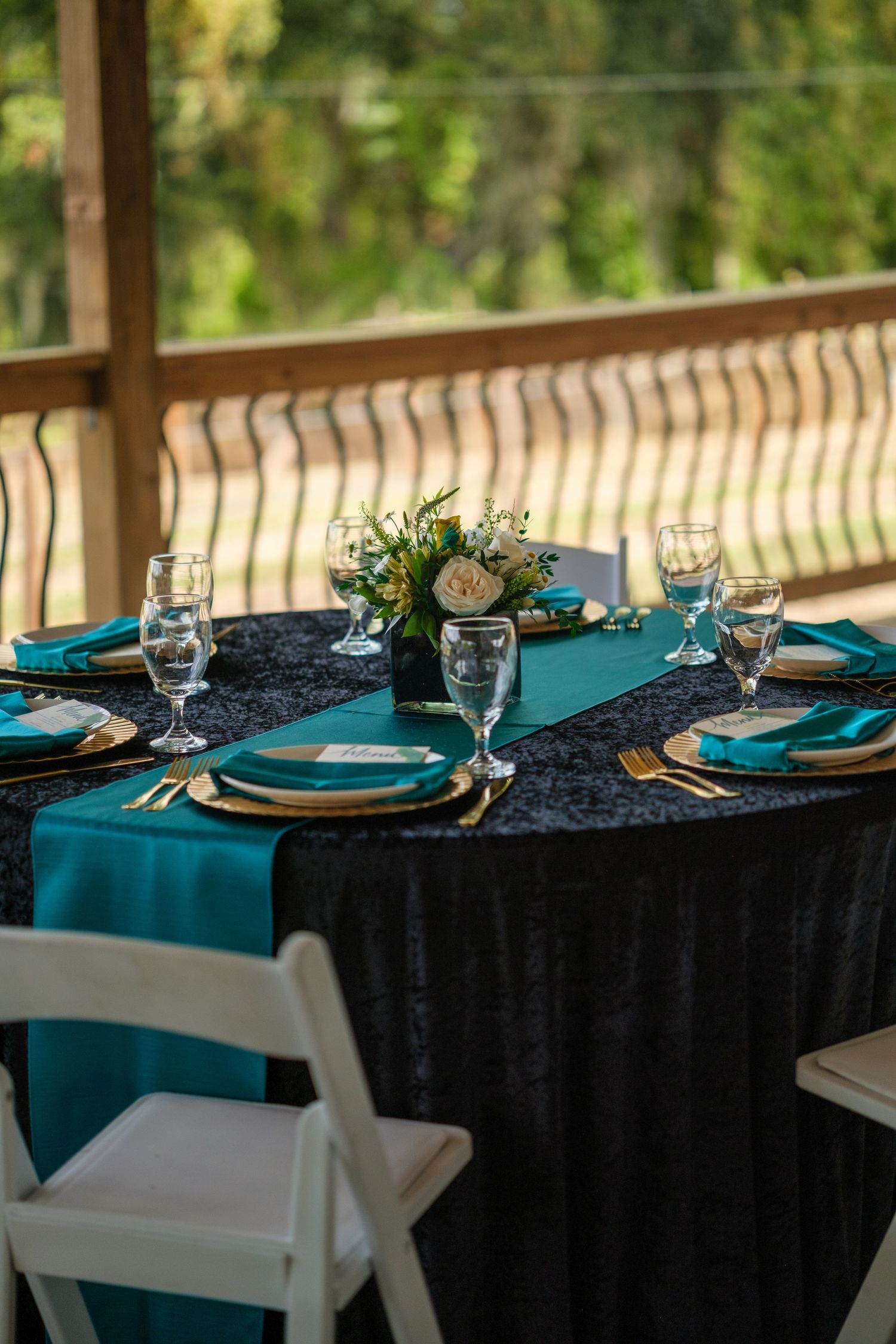 Elegant table setting with black tablecloth, teal napkins and white plates on a covered porch.