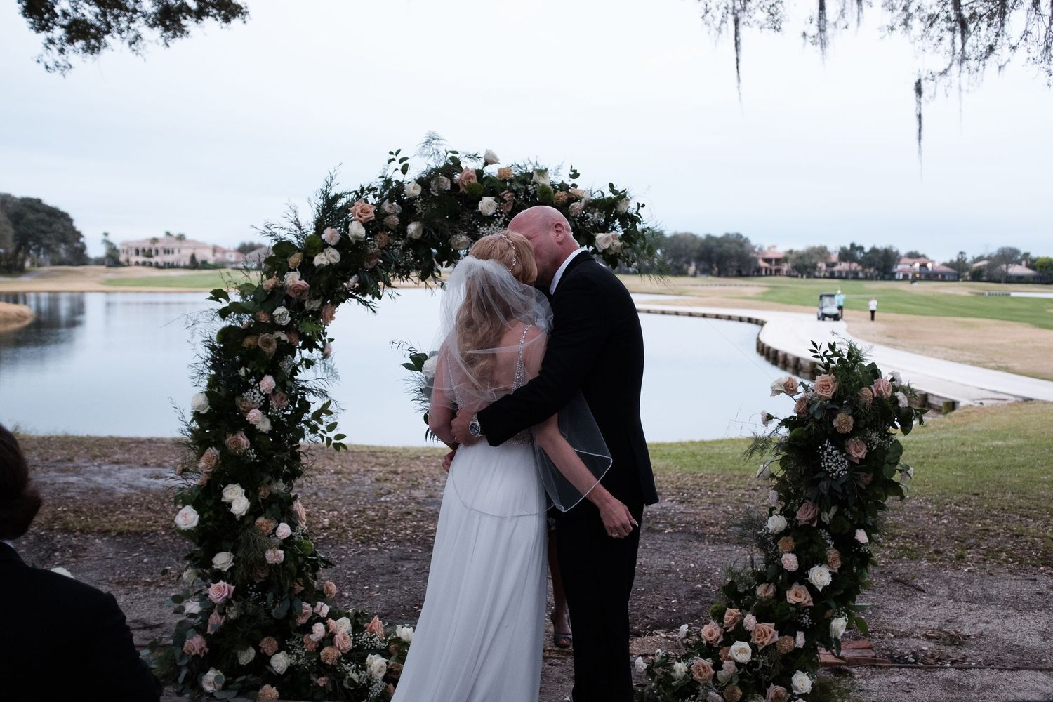Romantic wedding couple embraces under Spanish moss and floral archway at sunset by the lake.