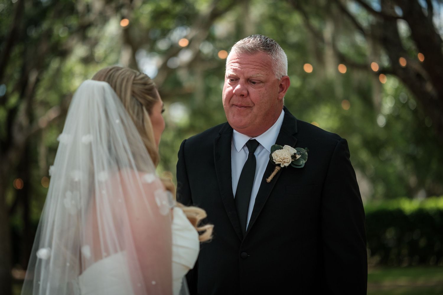 Groom in black suit with boutonniere smiling at bride in white veil during outdoor wedding ceremony.
