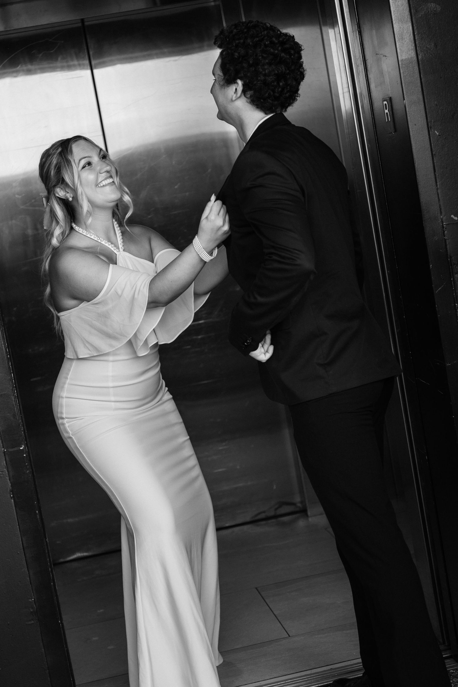 Black and white photo of a smiling bride in white gown with groom in black suit inside an elevator.