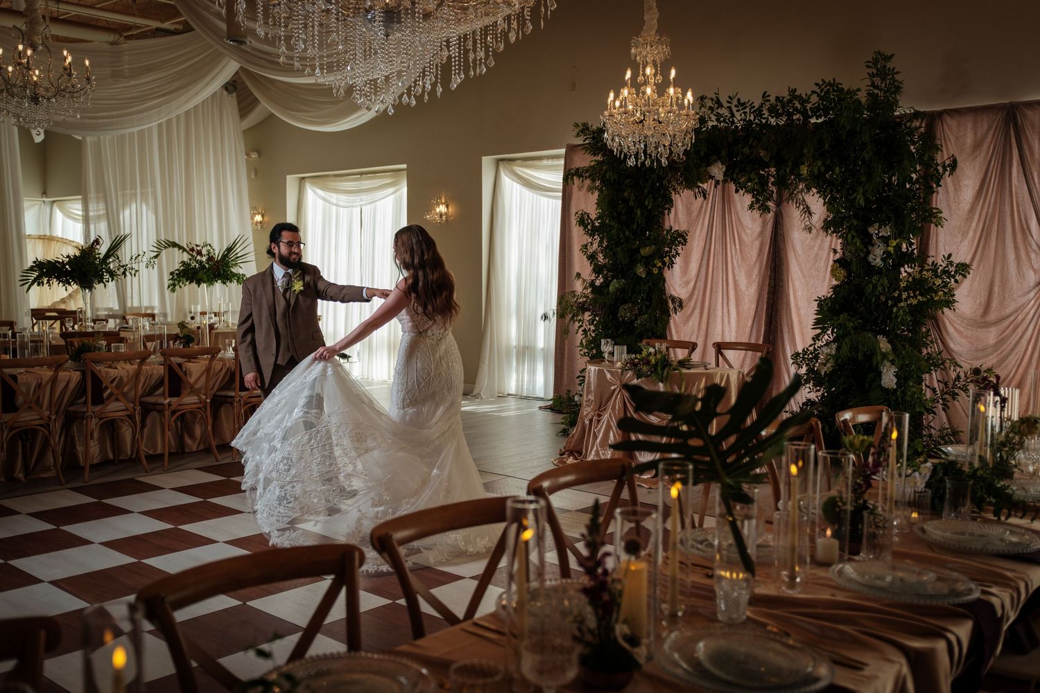 Bride and groom dancing in an elegant wedding reception hall with chandeliers, greenery arch, and candlelit tables.