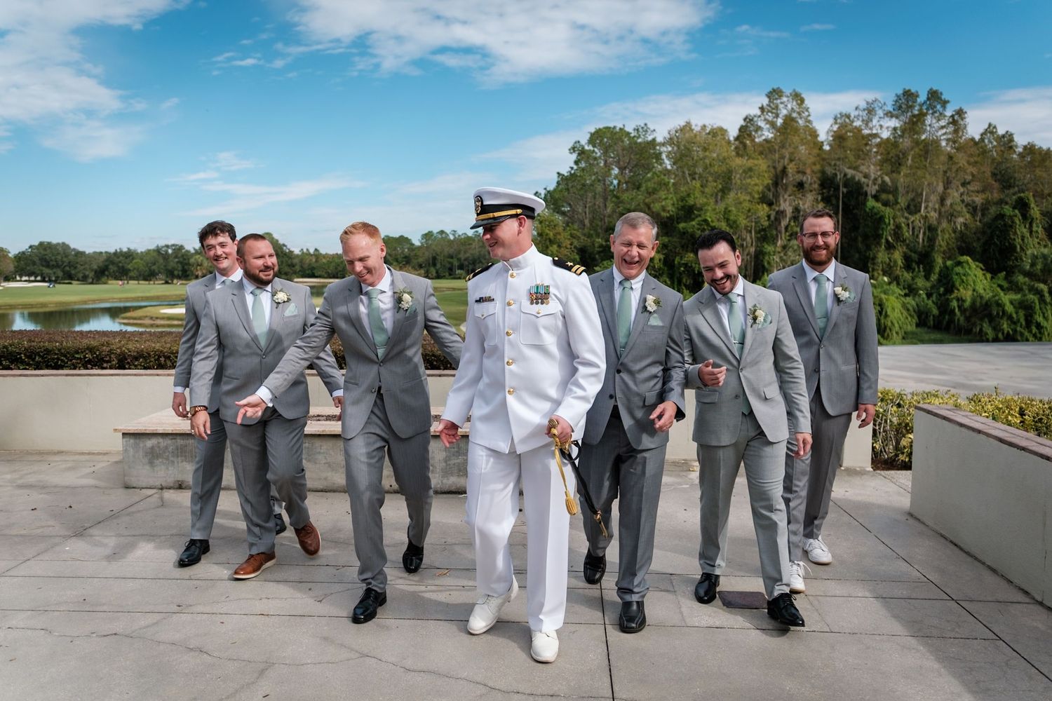 A group of groomsmen in gray suits walk alongside someone in white dress uniform on a sunny terrace overlooking water.