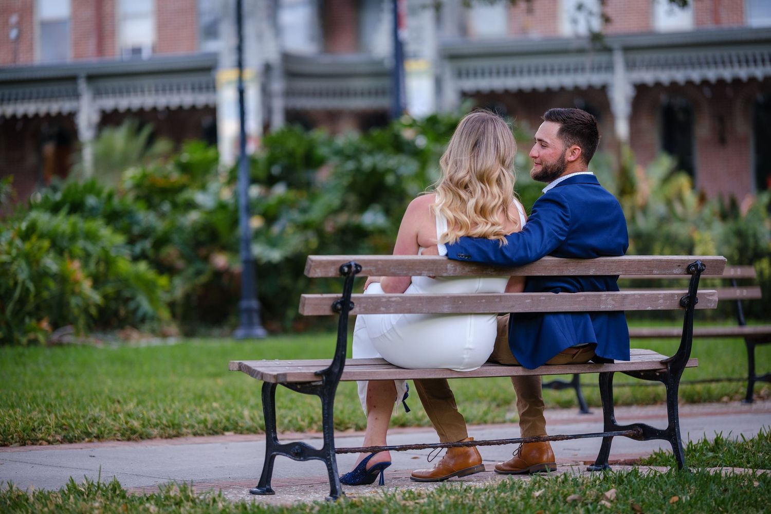 Bride and groom sitting together on a park bench, sharing a romantic moment in a lush green outdoor setting.