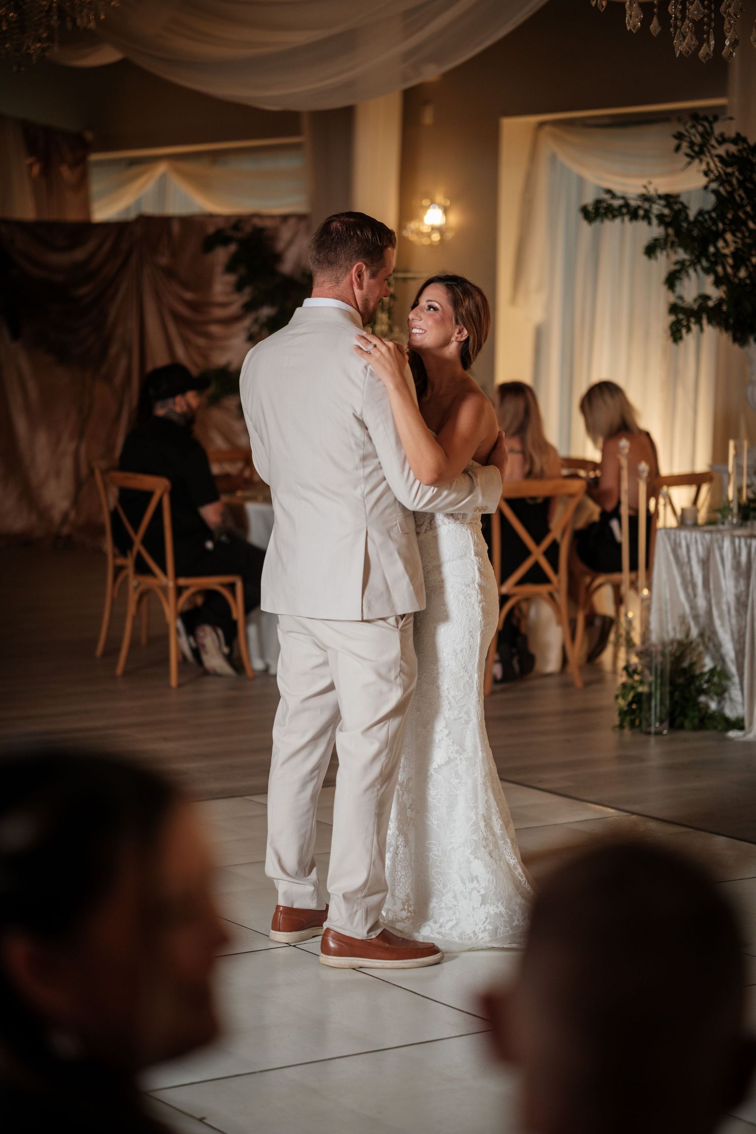 Bride and groom share their first dance at elegant wedding reception, bride in lace gown smiling up at groom in tan suit.