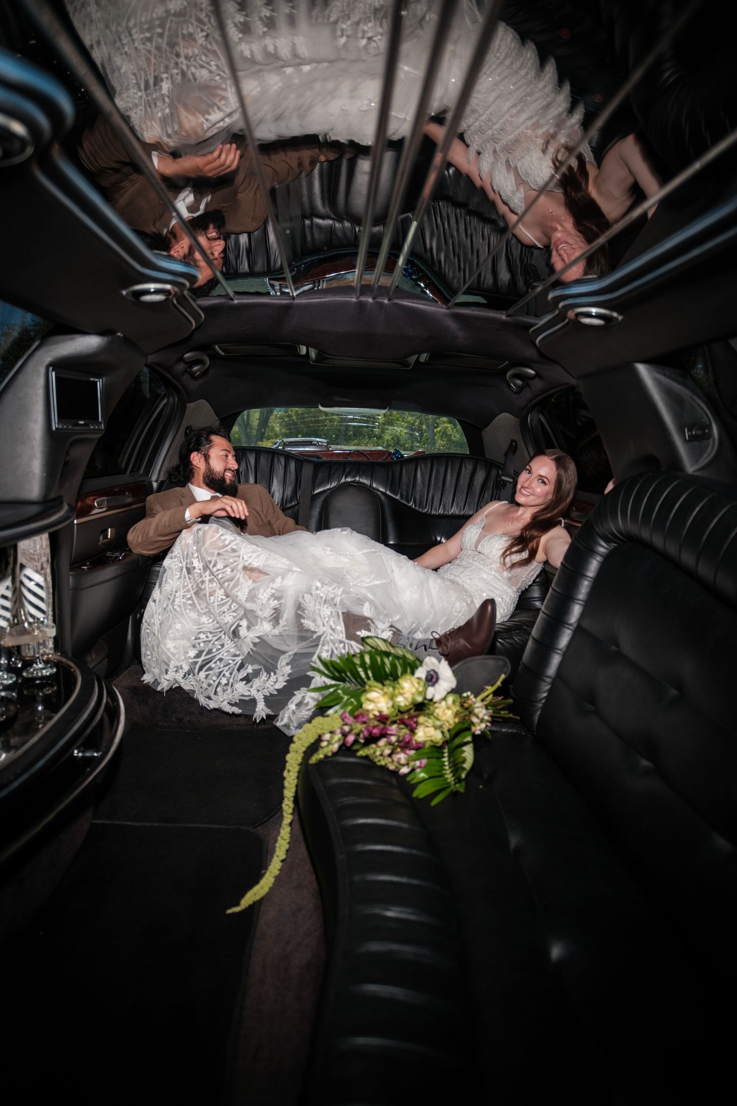 Bride and groom relaxing inside a luxurious black stretch limousine, with bridal bouquet on the seat.