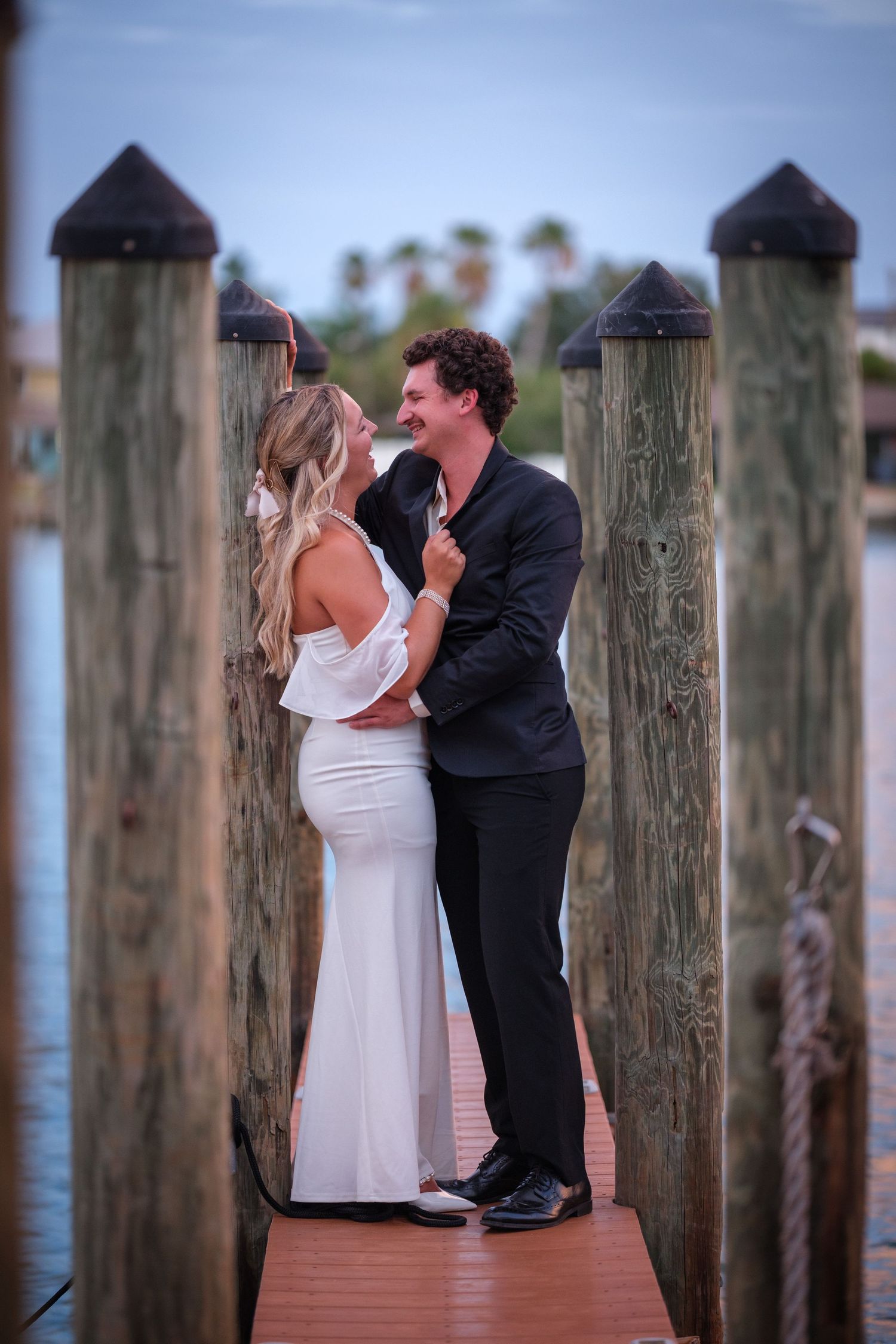 Couple in formal wear sharing a romantic moment on a wooden dock by the water at dusk.