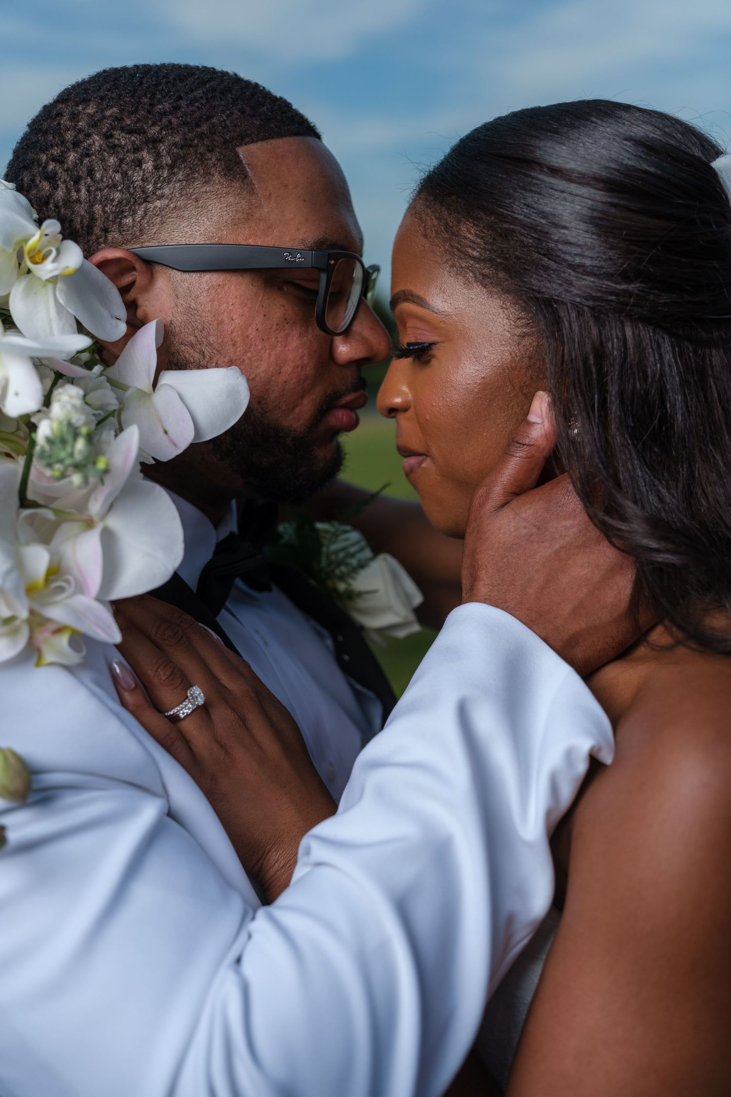 A Black couple shares an intimate moment in wedding attire, bride holding white orchids, groom tenderly cupping her face.