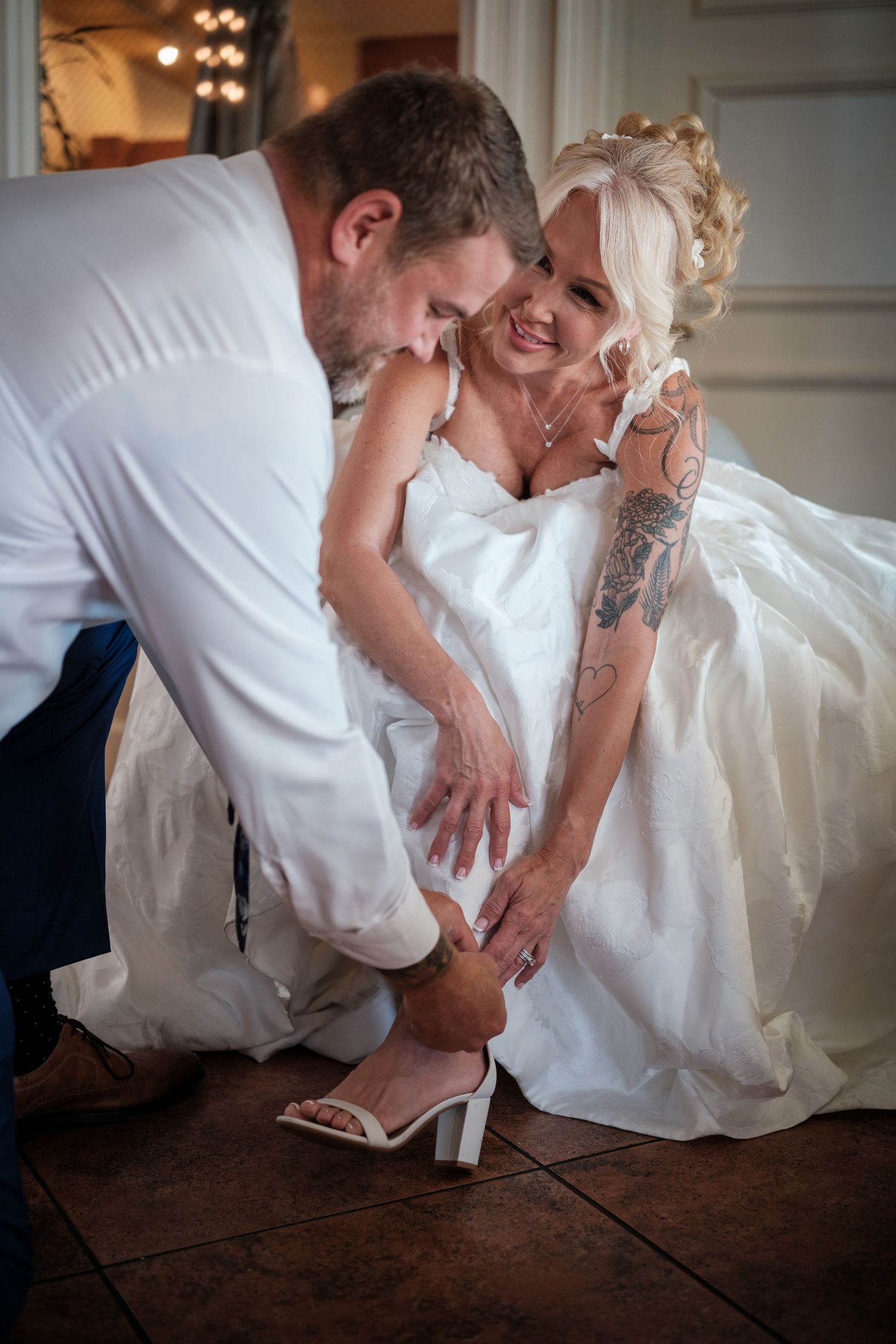 Groom kneeling to put wedding shoe on tattooed bride in white gown, sharing a tender romantic moment.