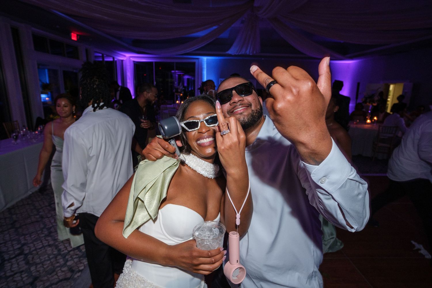 Joyful couple in white outfits celebrating at a wedding reception with purple ambient lighting.