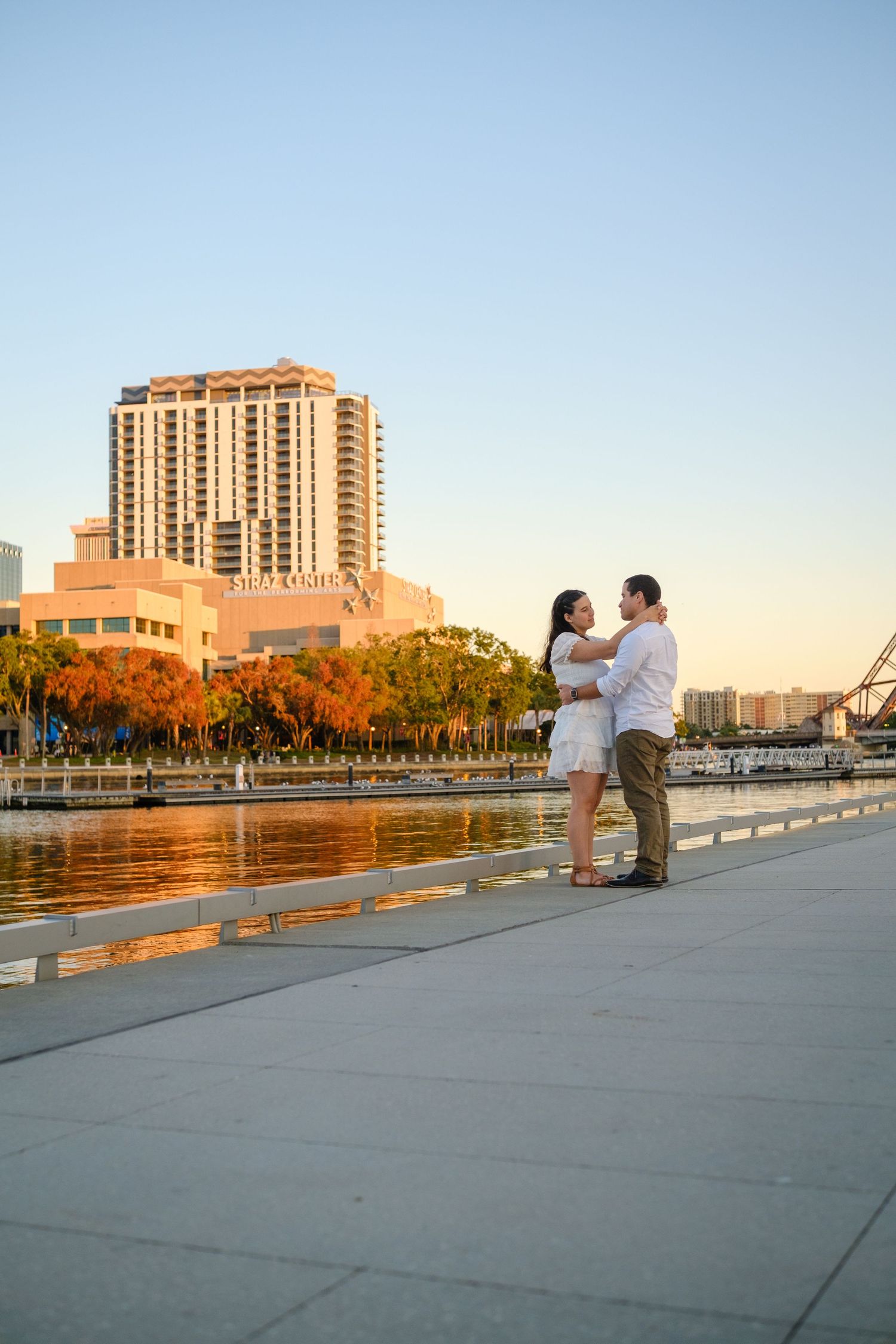 Couple embracing at golden hour by Tampa waterfront with city skyline and reflection pool in background.