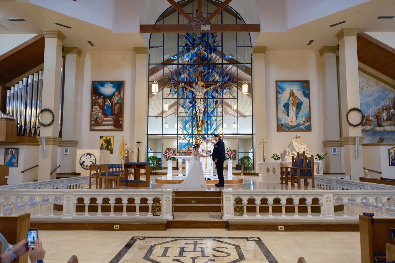 Couple exchanging vows at ornate Catholic church altar with stunning stained glass windows and IHS mosaic floor.