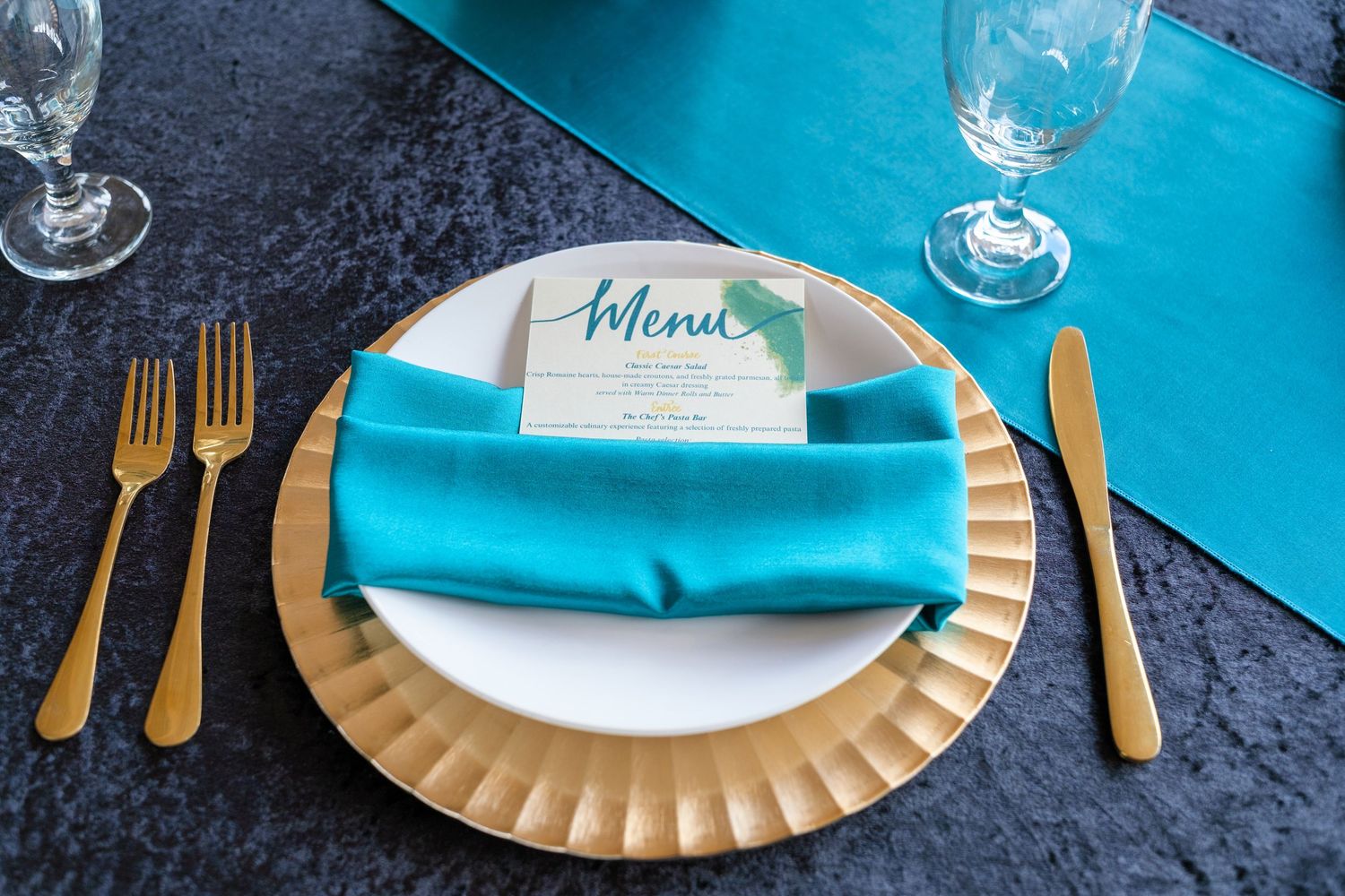 Elegant place setting with turquoise napkins, gold flatware, and menu card on textured charger plate.