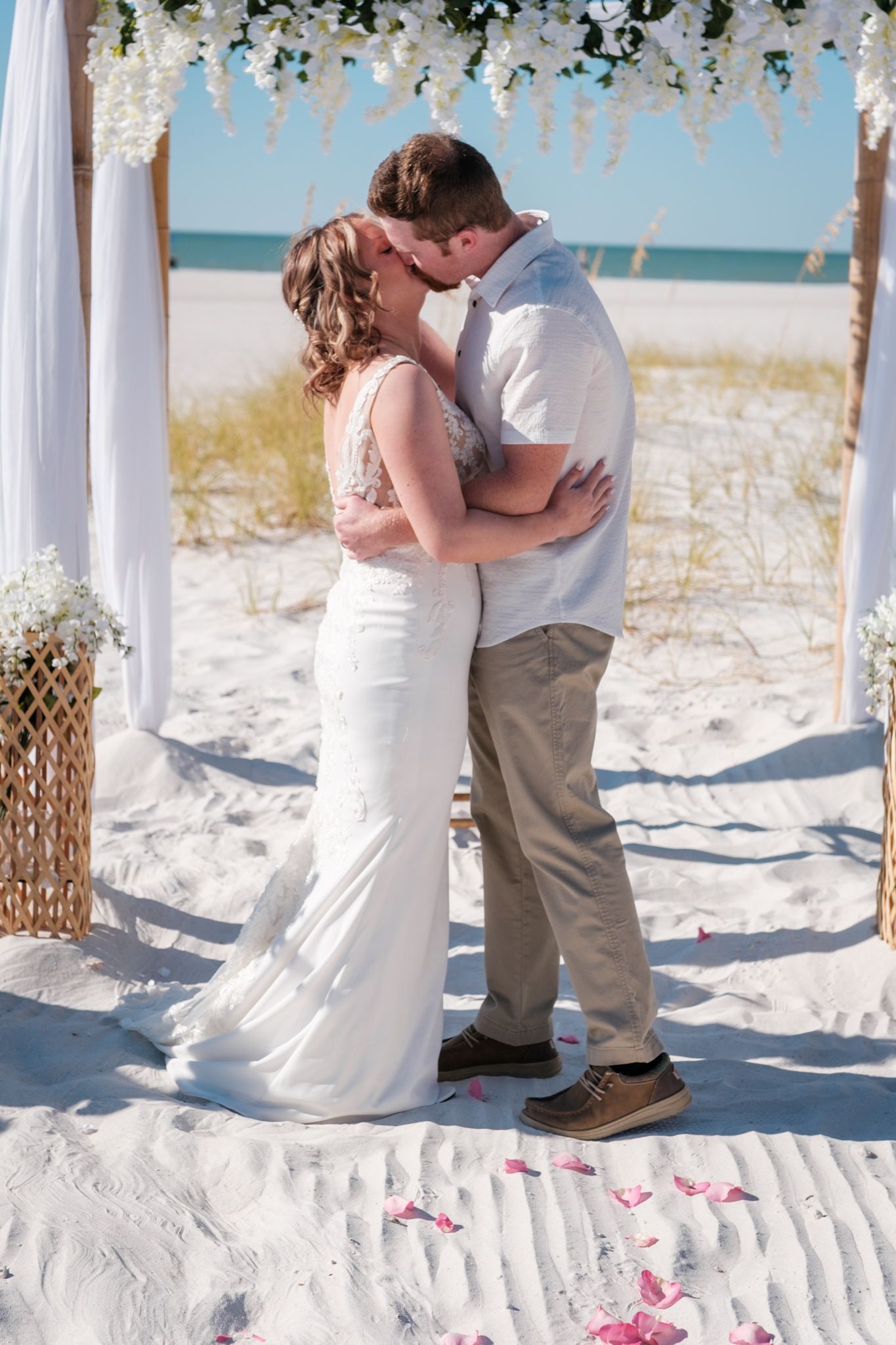 Romantic beach wedding ceremony moment under floral-decorated white arch with ocean backdrop.