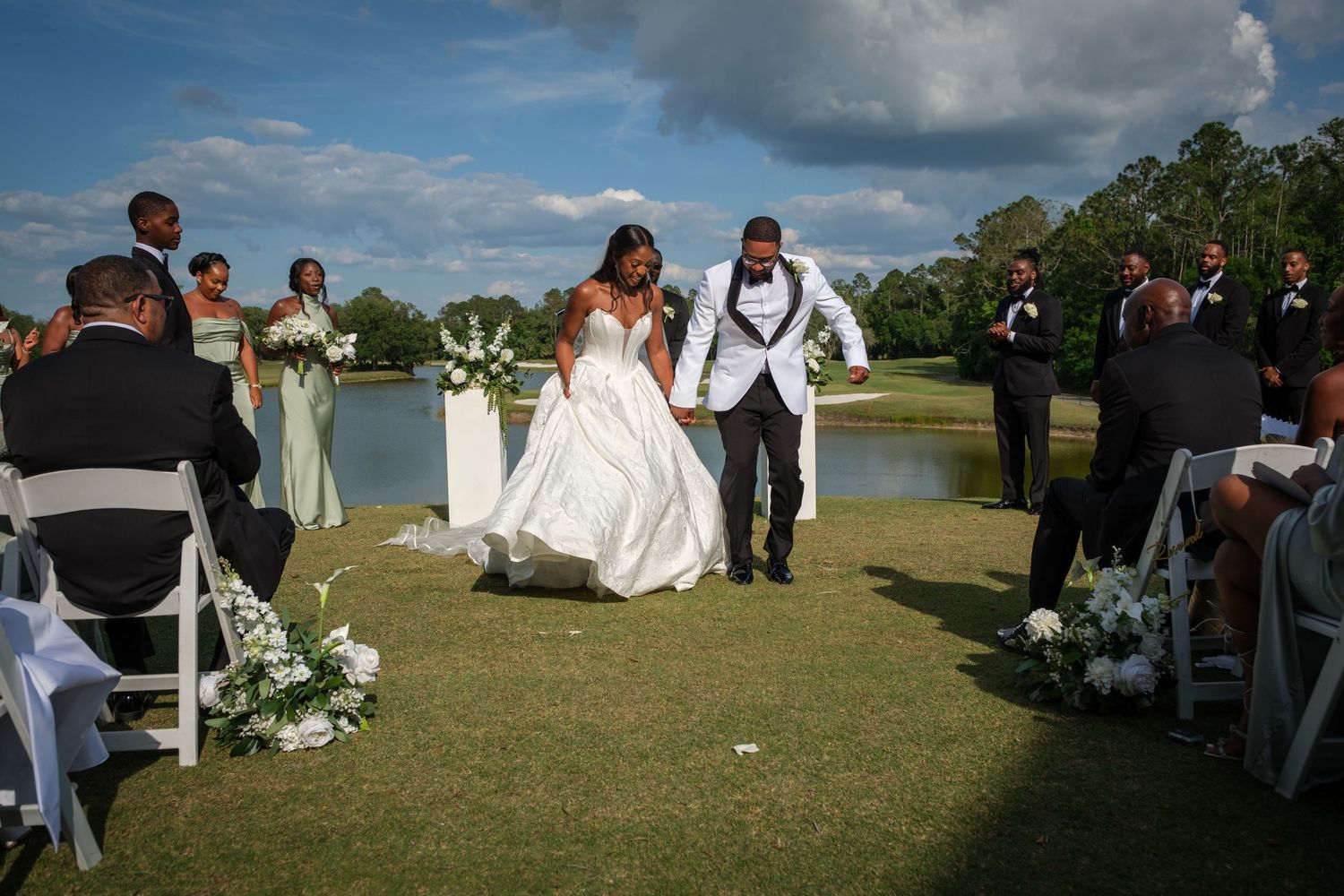 Bride and groom walk down outdoor aisle at golf course wedding ceremony with bridal party and white chairs.