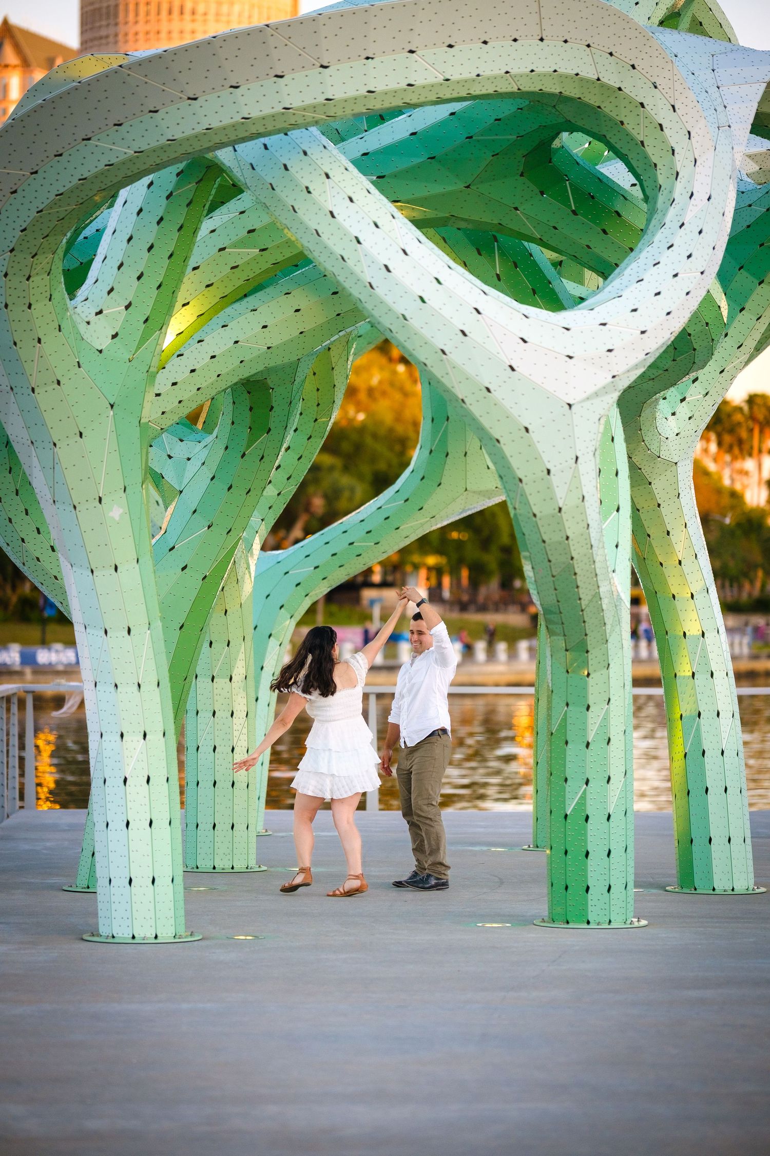 Couple dancing under a large green abstract sculpture near a waterfront at golden hour.