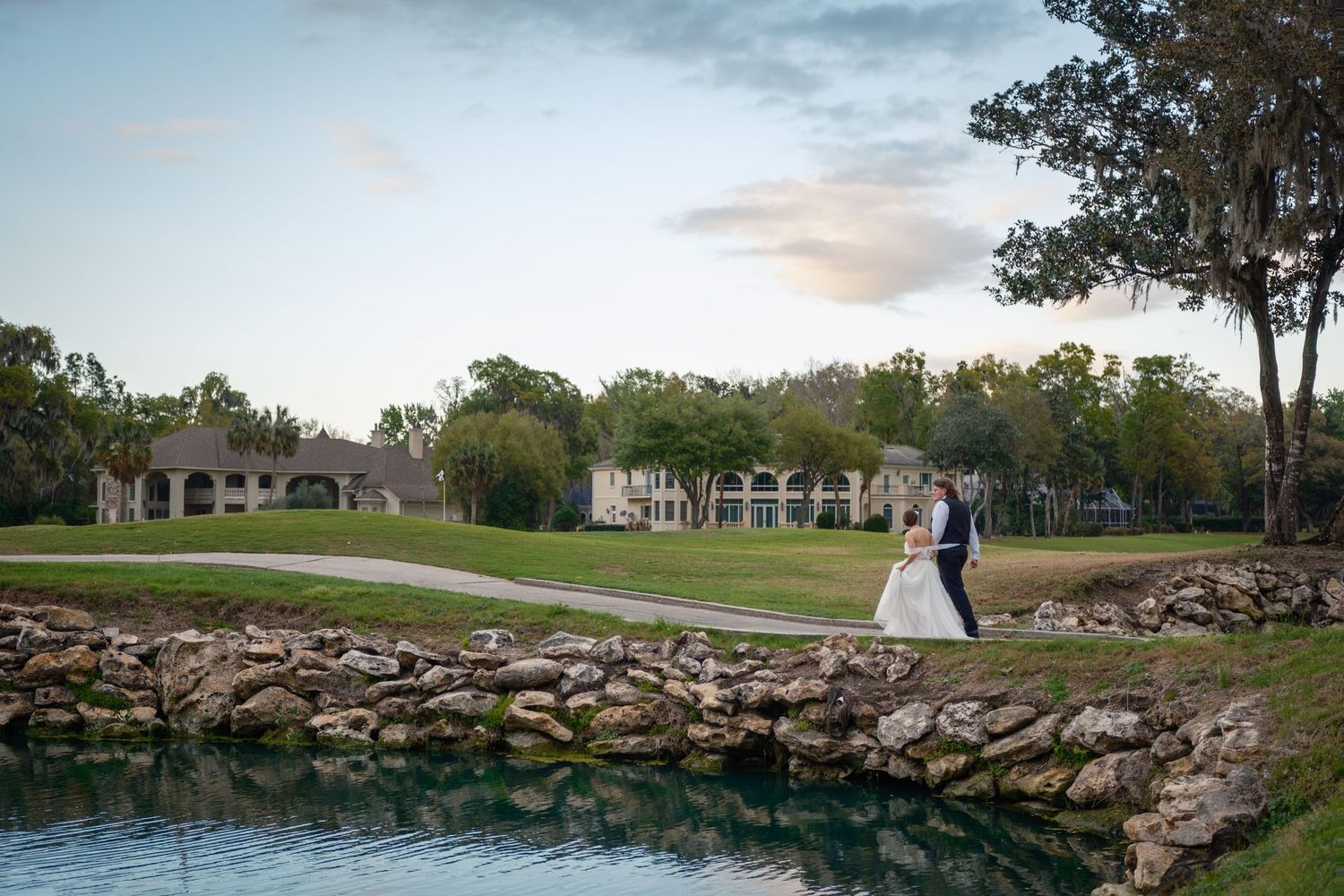 Bride and groom share a romantic moment on a stone bridge over a pond at a lush golf course wedding venue.