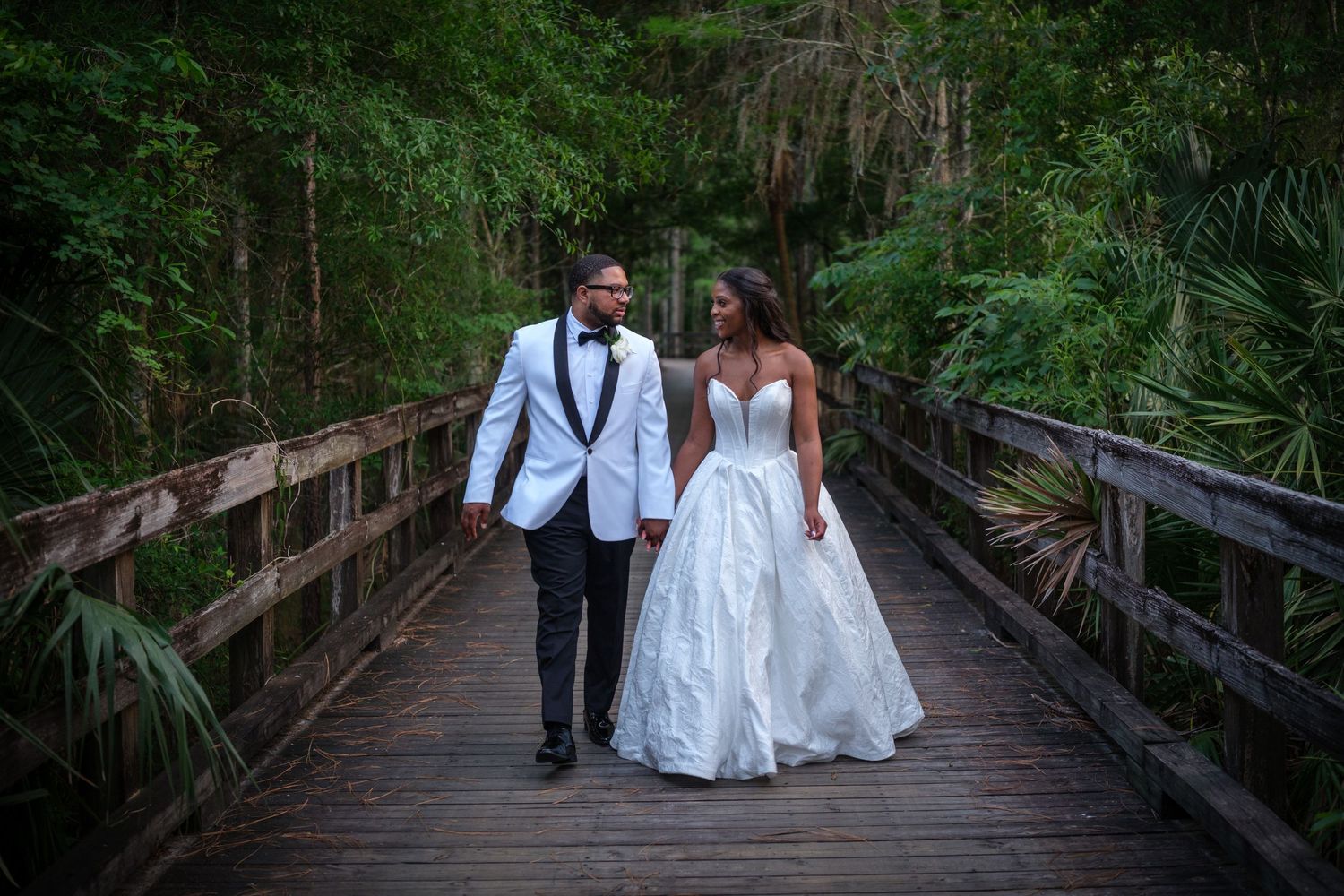 Bride and groom walk hand-in-hand on a wooden bridge surrounded by lush green forest on their wedding day.