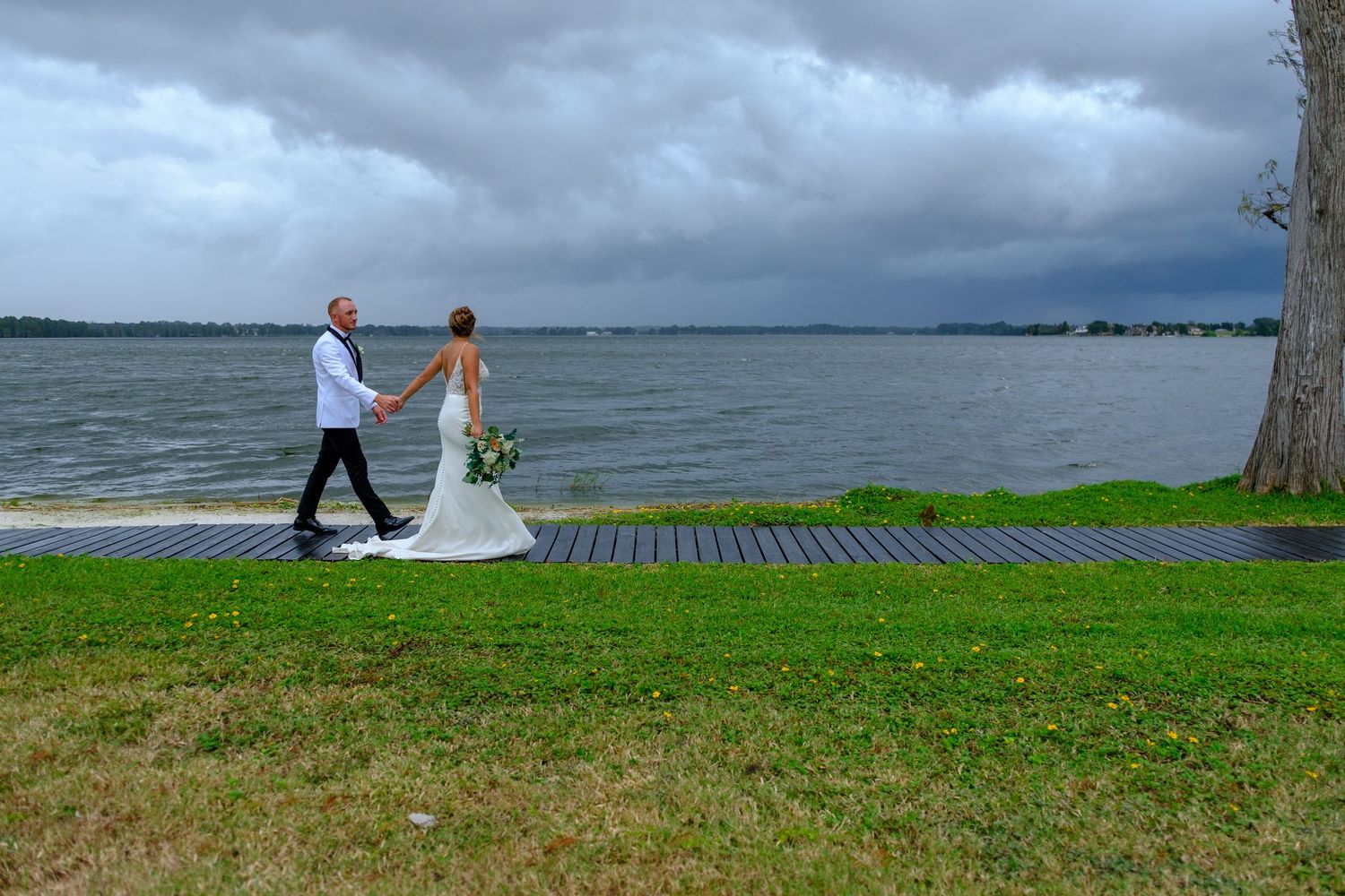 A couple in wedding attire walks hand in hand along a wooden dock by a stormy lake with dark clouds overhead.
