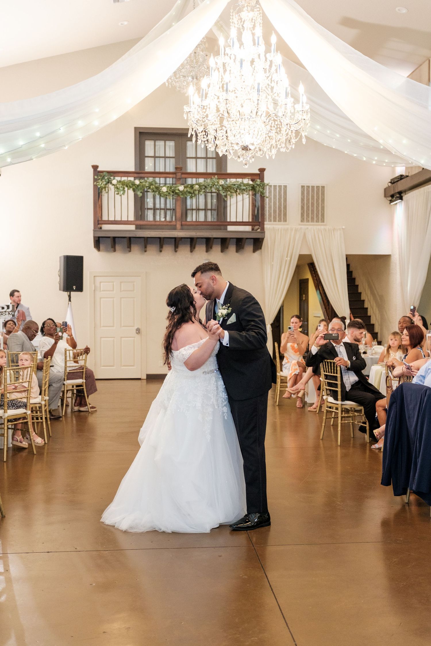 Couple sharing their first dance in an elegant white-draped ballroom with crystal chandeliers and watching guests.