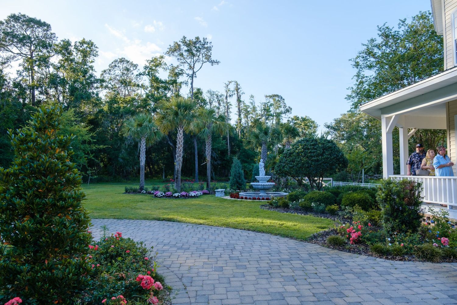 A curved brick paver walkway leads through a lush garden with a fountain and Spanish moss draped trees in the background.