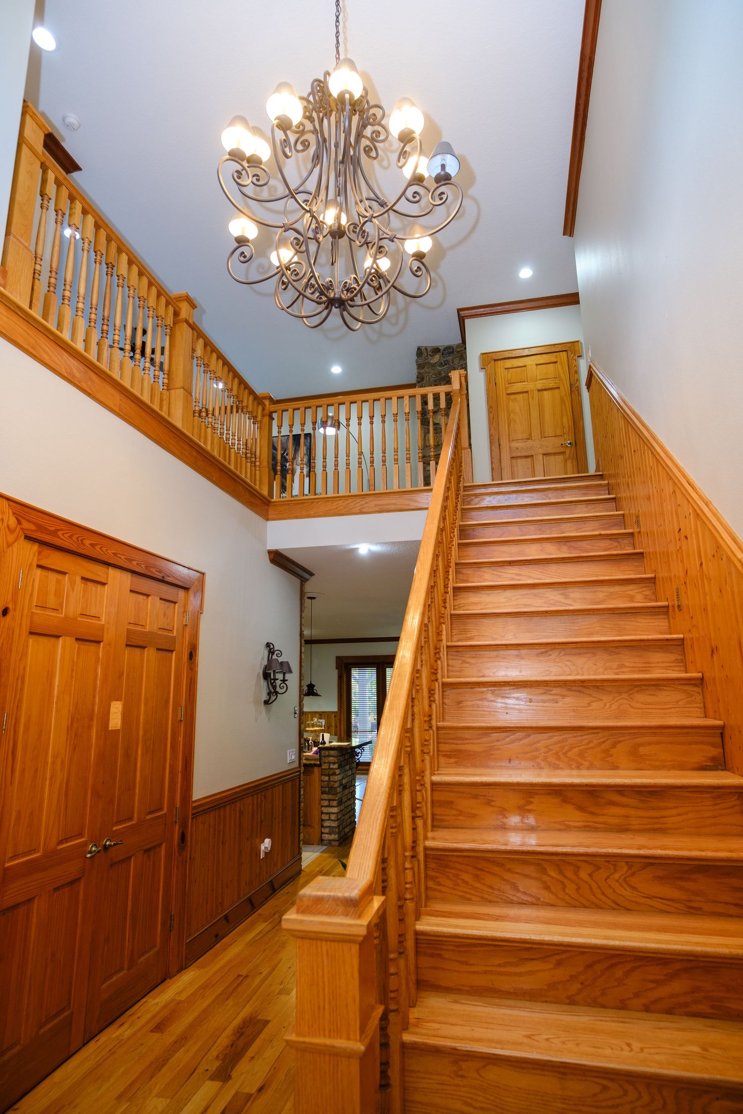 Elegant wood staircase and entryway with ornate chandelier features oak railings and warm honey-toned hardwood floors.