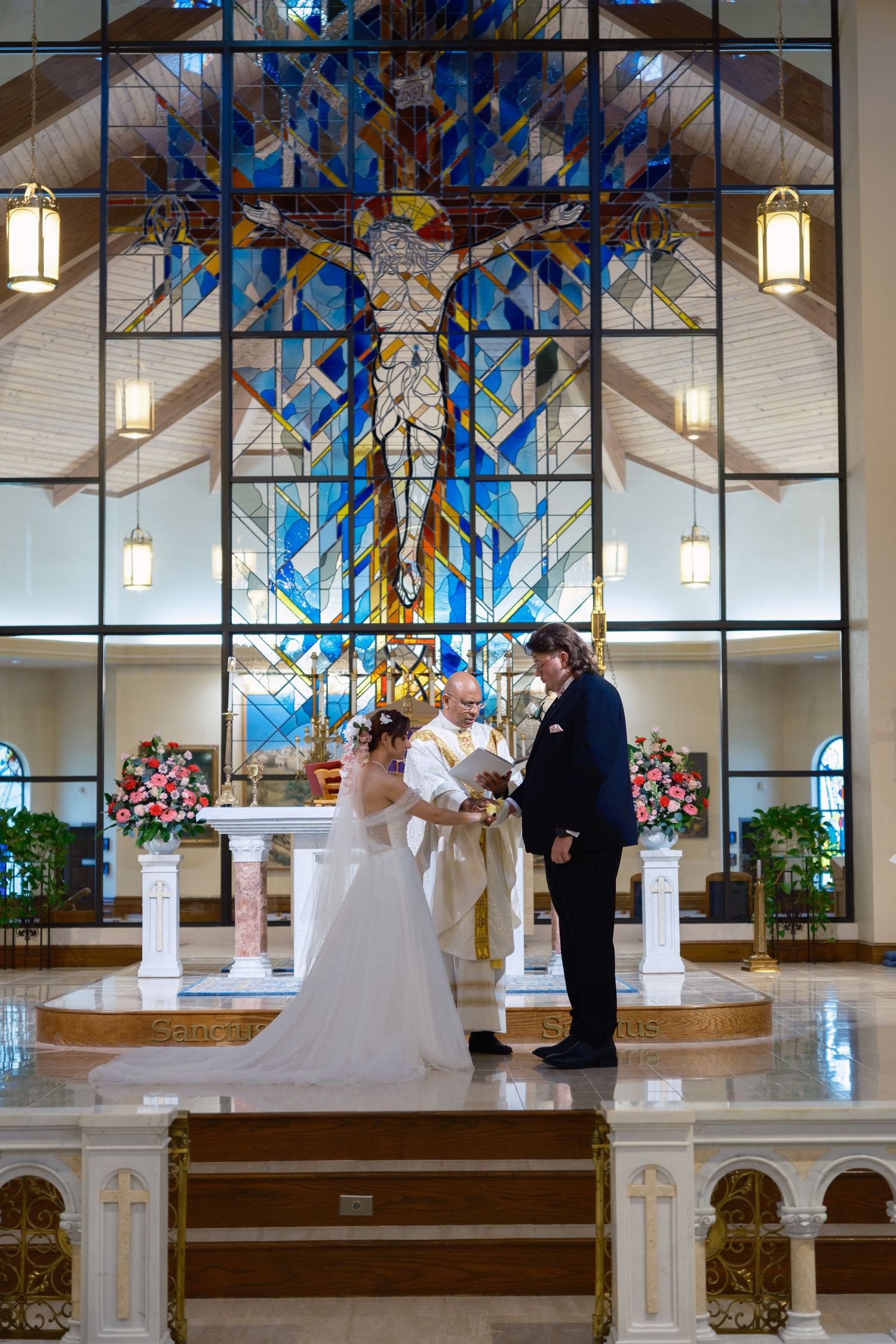Bride and groom exchange vows at church altar with stunning blue stained glass window backdrop during wedding ceremony.