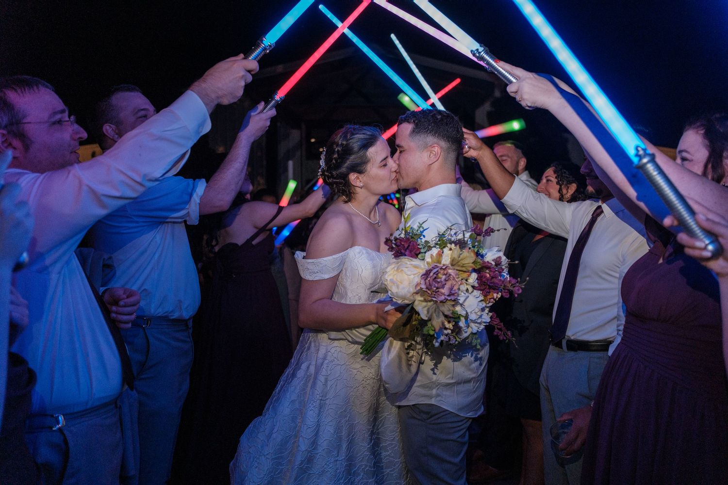 Newlyweds share a kiss surrounded by guests holding colorful lightsabers at night.