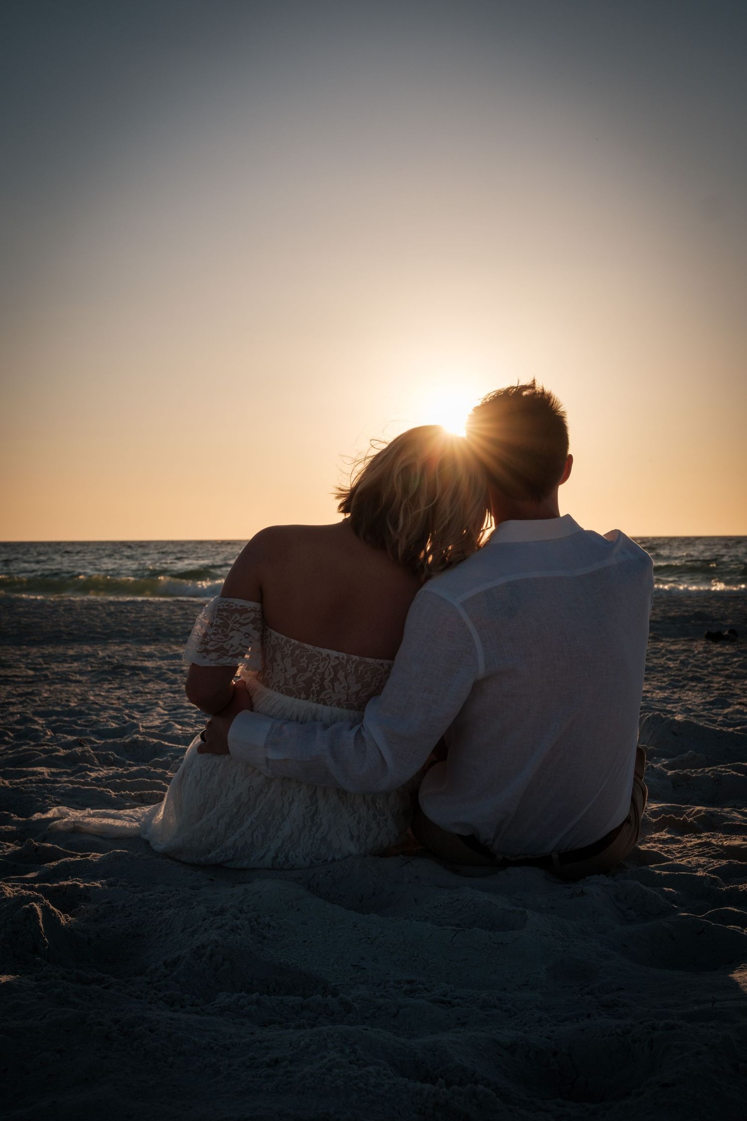 Couple sitting together on beach at sunset, silhouetted against golden sky, sharing a romantic embrace by the ocean.