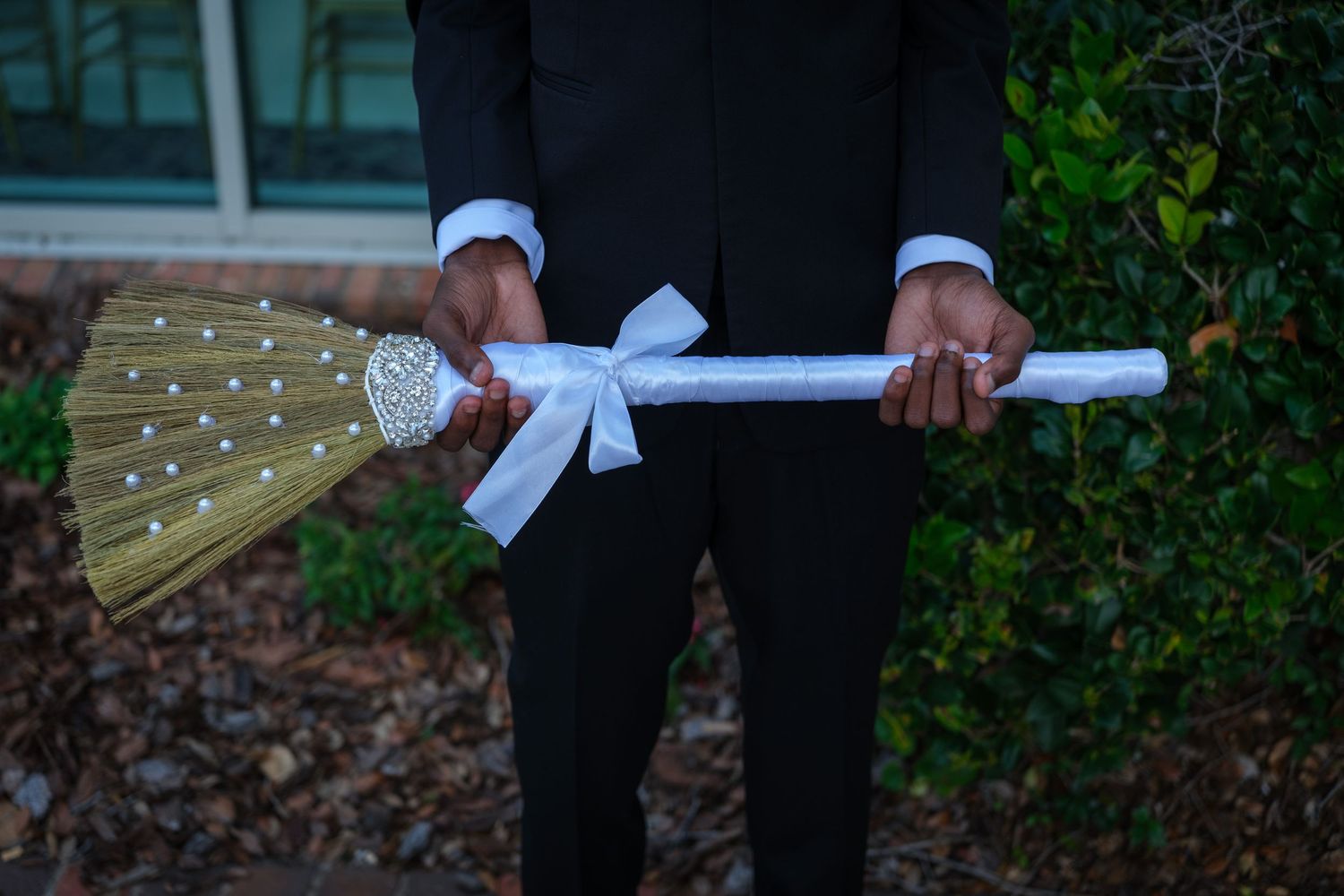 Person in black suit holding a decorative wedding broom with white bow and rhinestone details for jumping the broom ceremony.