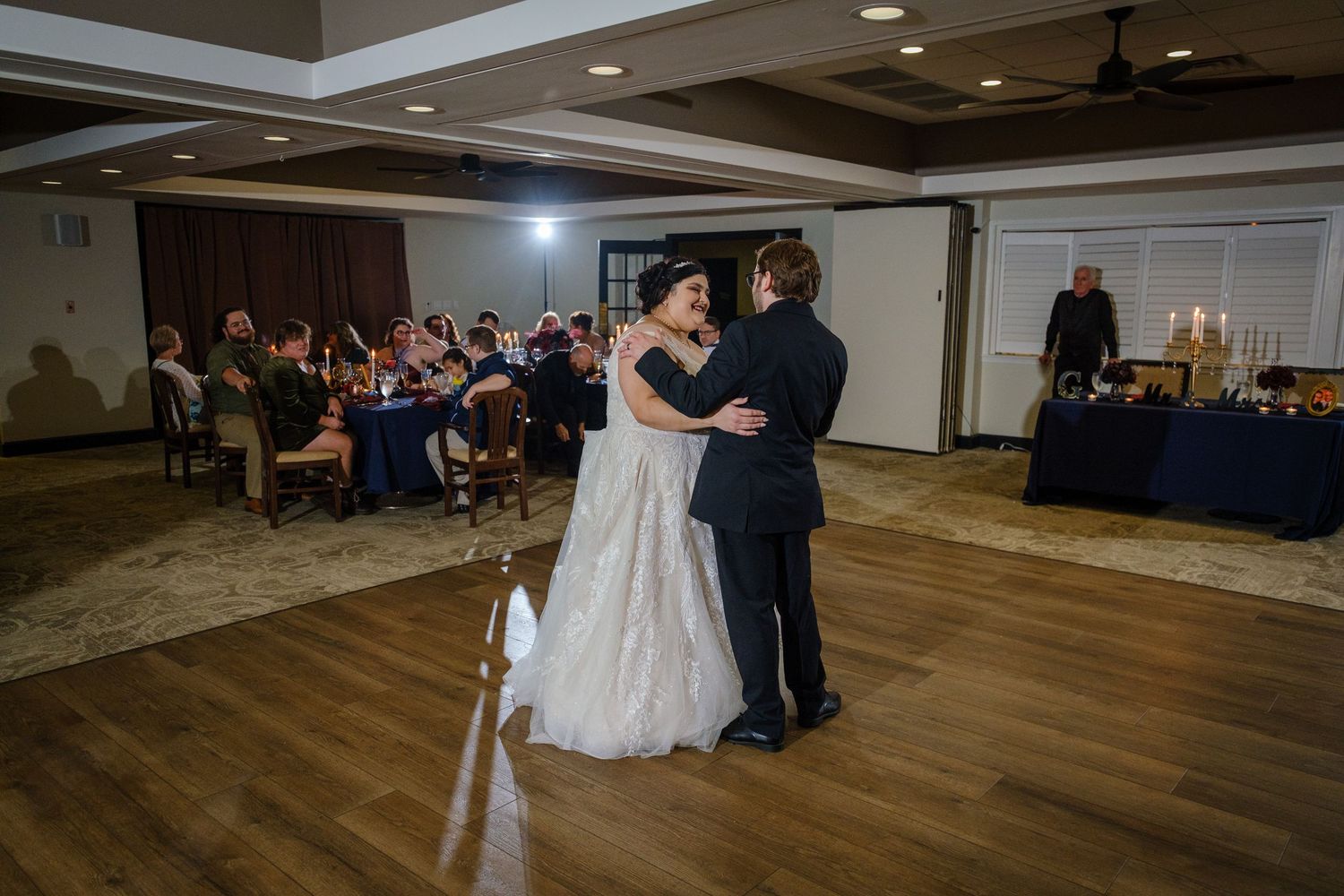 A couple shares a romantic dance together in a spacious ballroom with wooden floors and recessed lighting.