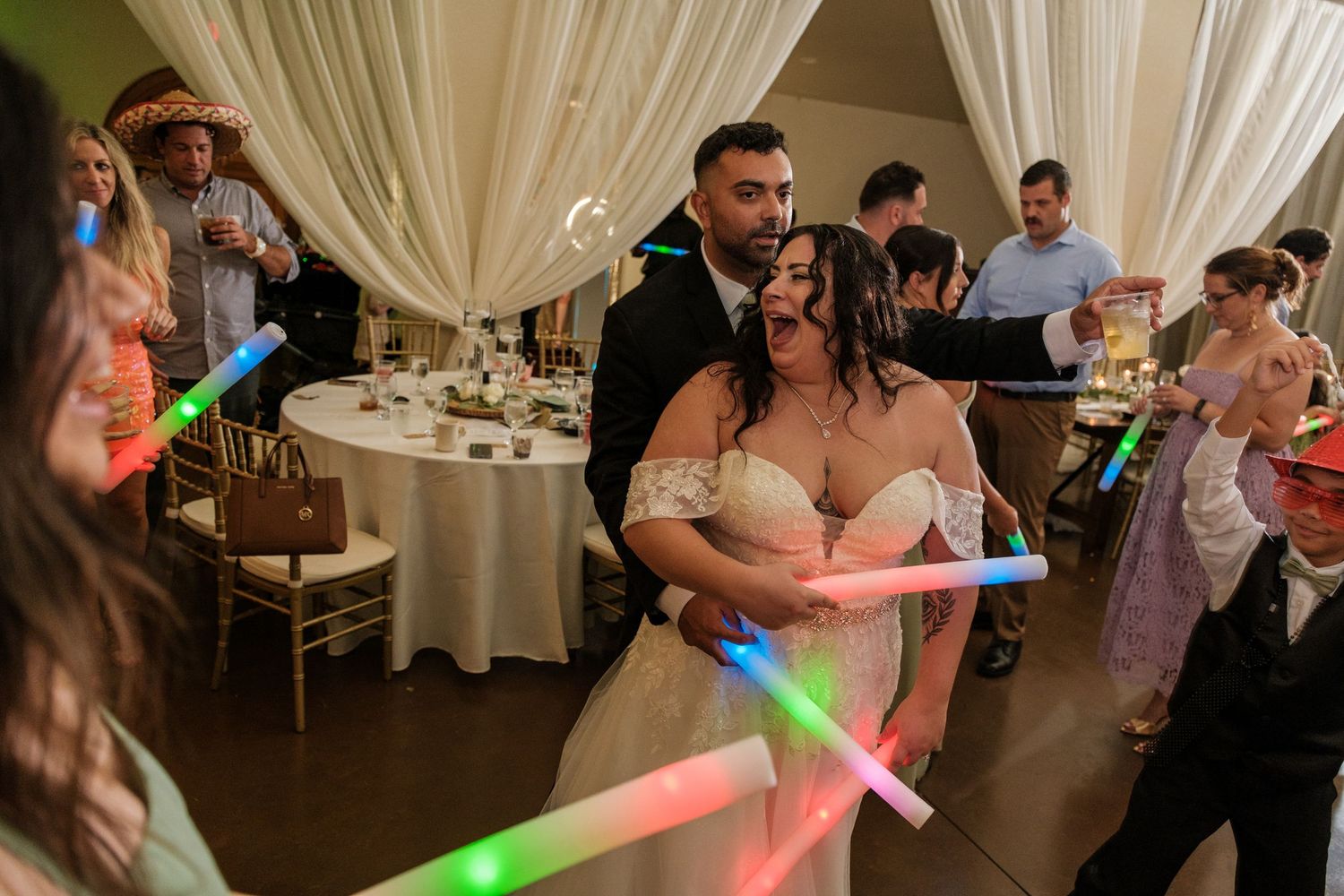 Wedding guests dance with colorful LED foam glow sticks on a dimly lit dance floor at a reception celebration.