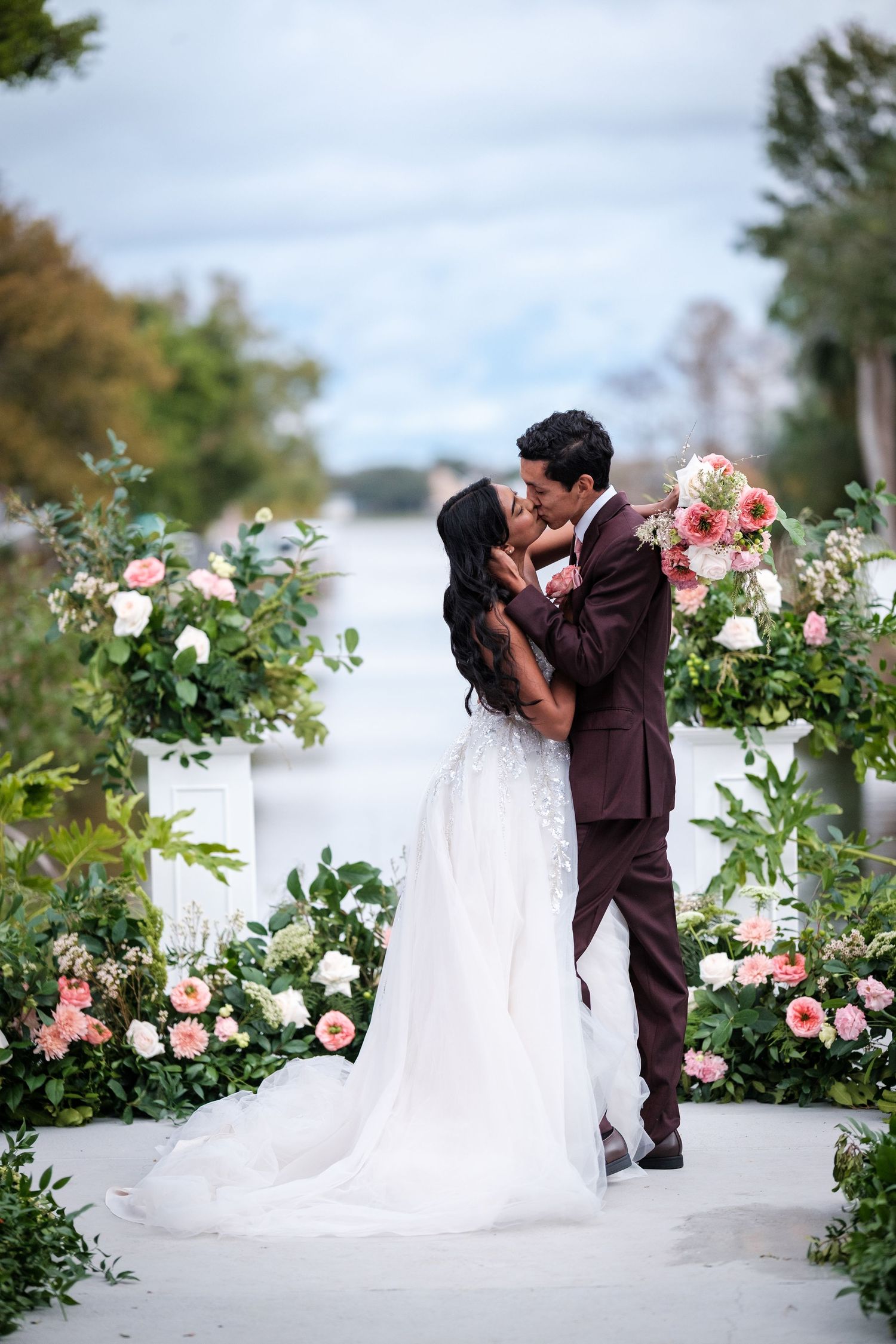 A romantic wedding couple embraces in a garden setting surrounded by pink and white floral arrangements.