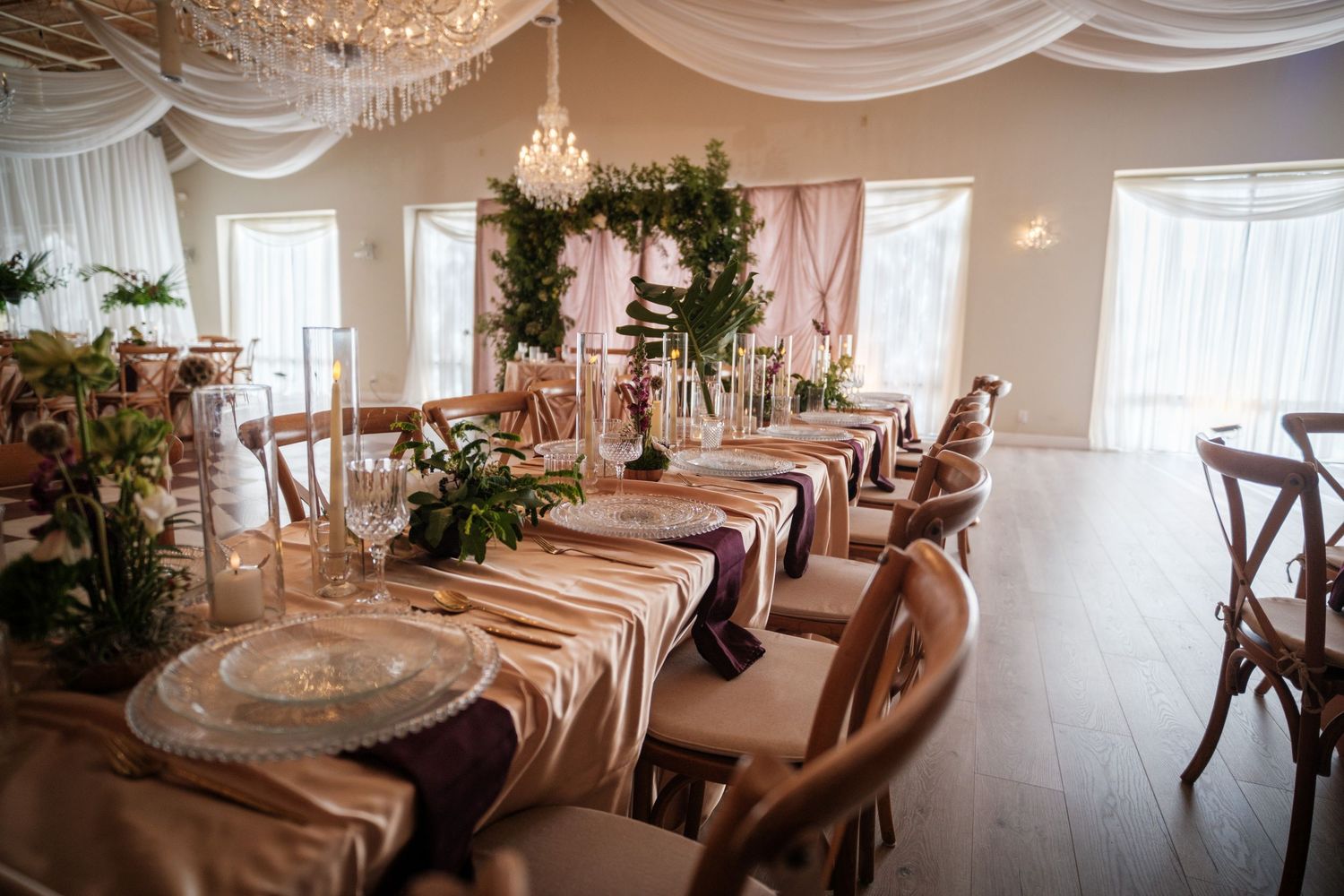 Elegant wedding reception hall with long tables, gold linens, floral centerpieces, and cross-back chairs in a draped venue.