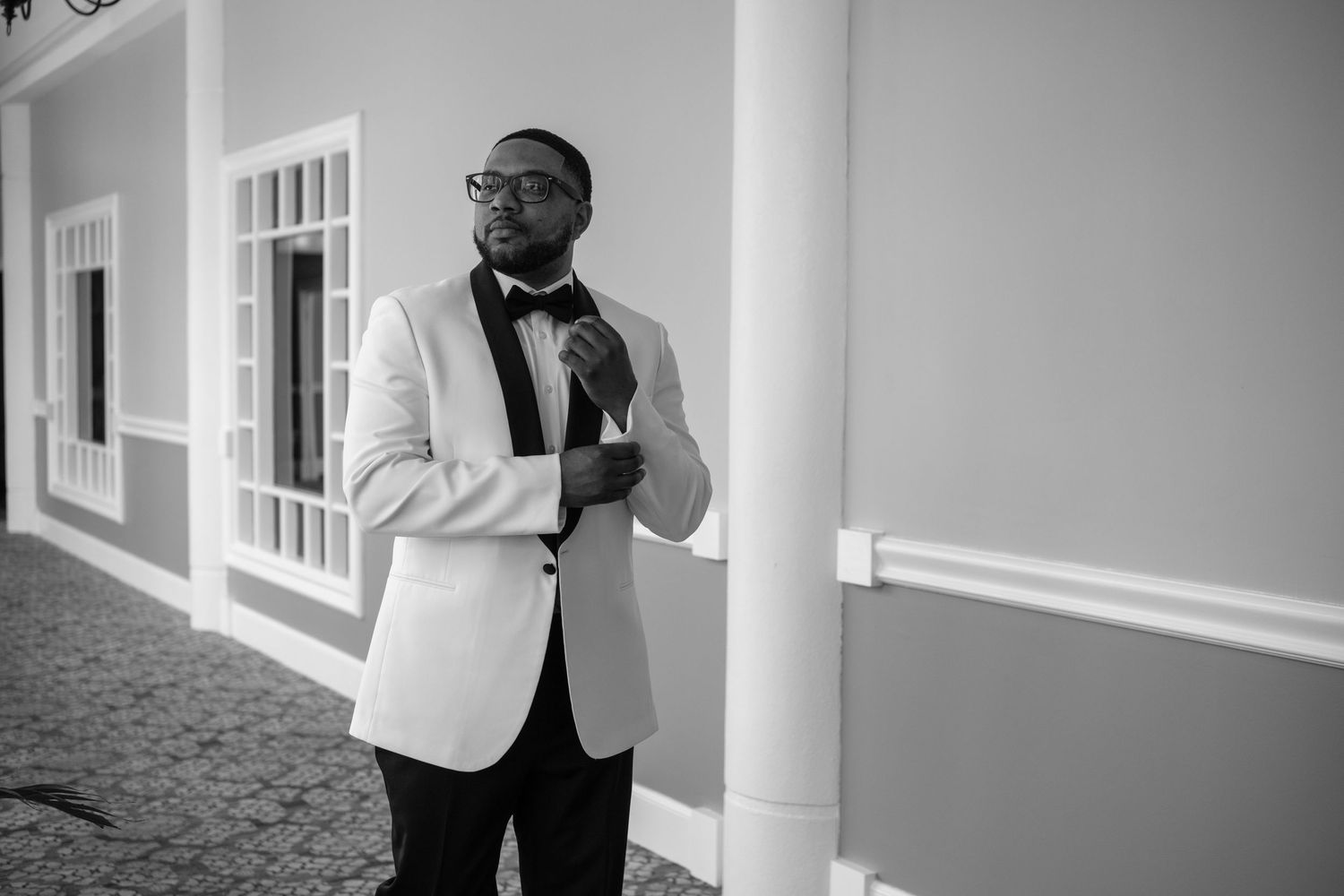 A dapper groom in a white tuxedo jacket adjusts his bow tie outside an elegant building in a black and white photo.
